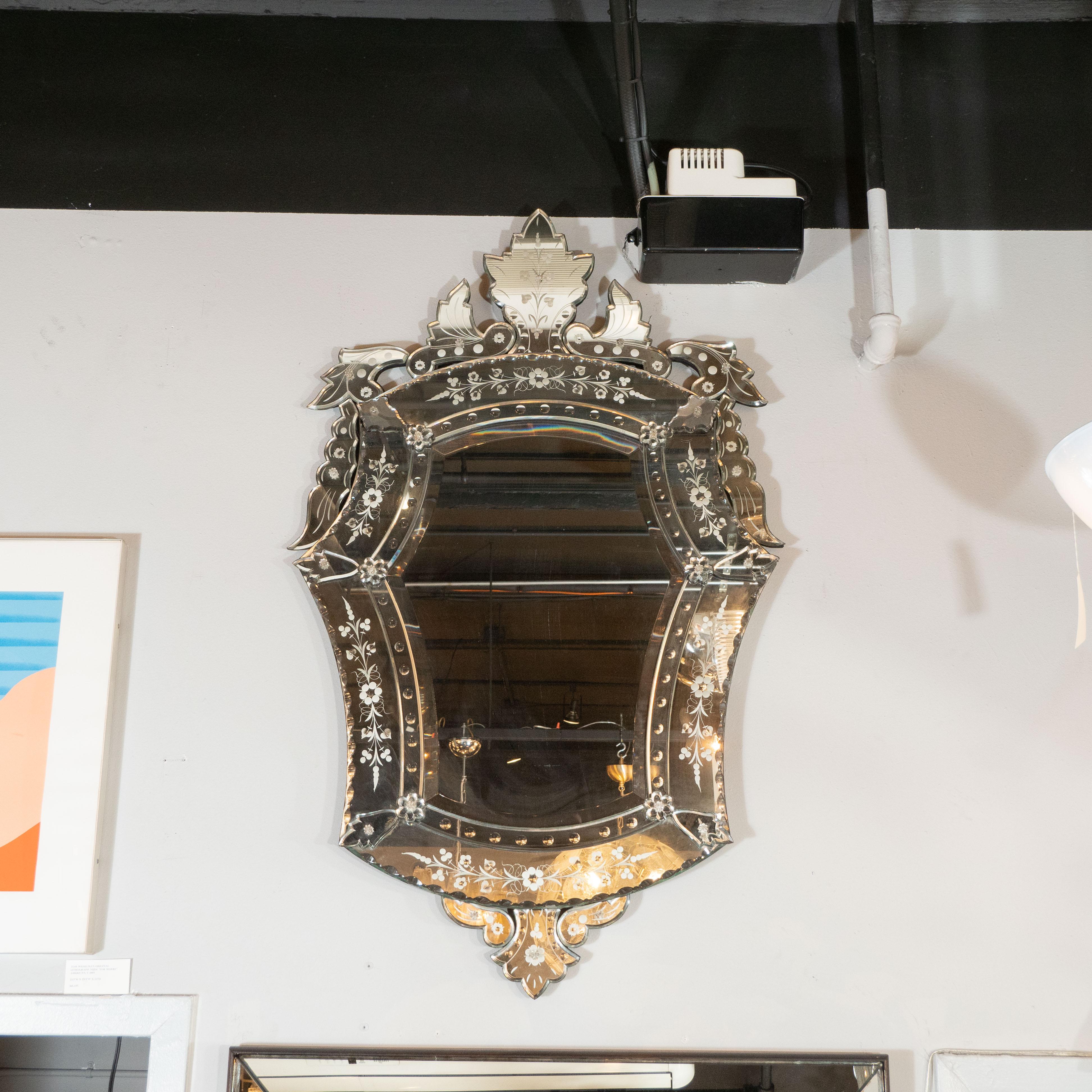 This glamorous mirror was realized in Italy, circa 1950. It offers a cartouche form with mirrored forms resembling stylized leaves adorning the perimeter of the piece. The mirror offers etched foliate detailing throughout as well chain and bevelled