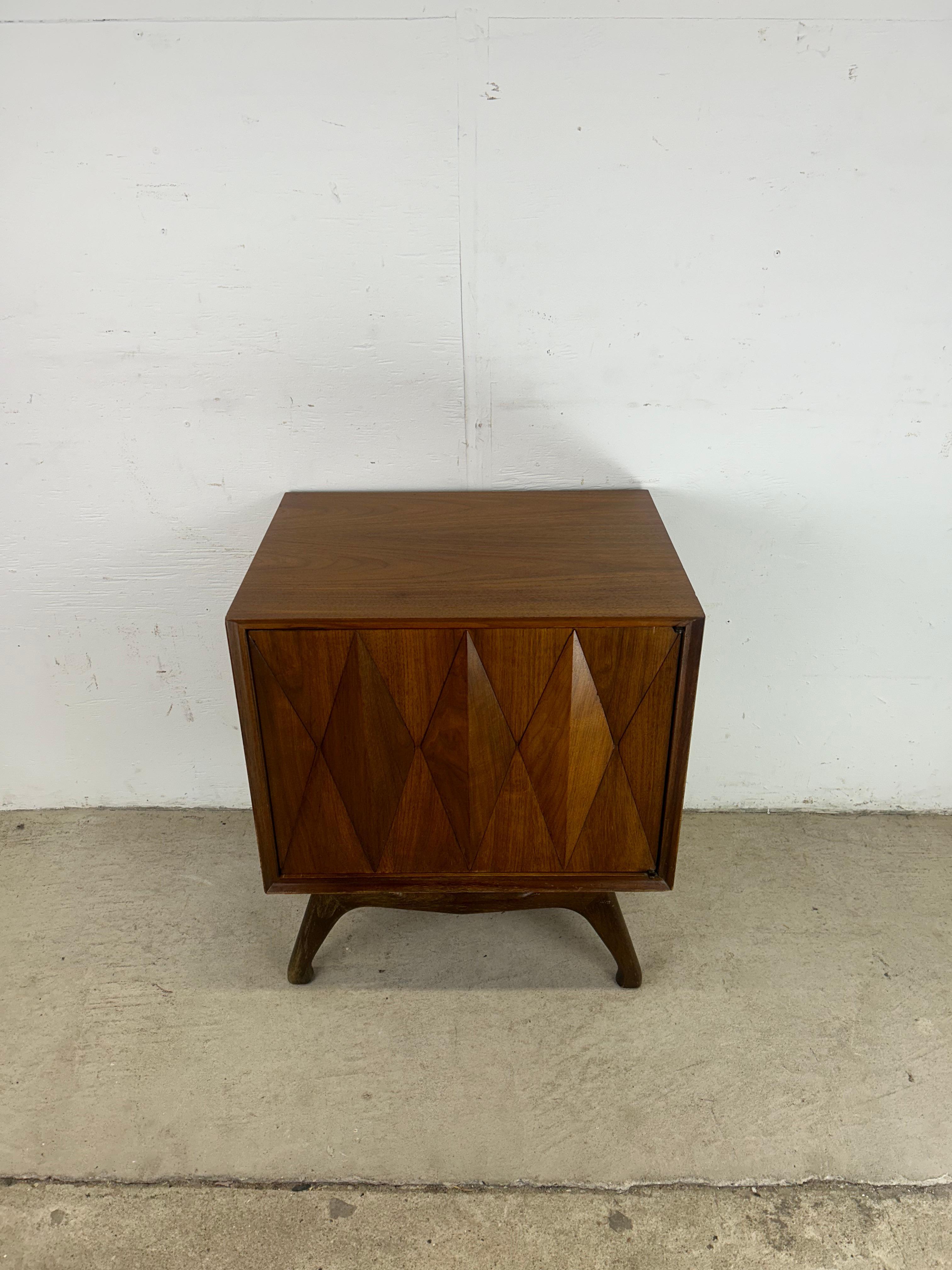 This mid century modern nightstand by Albert Parvin for American of Martinsville features hardwood construction, walnut veneer with original finish, single cabinet with unique diamond pattern on door, interior shelf, and tapered legs with a