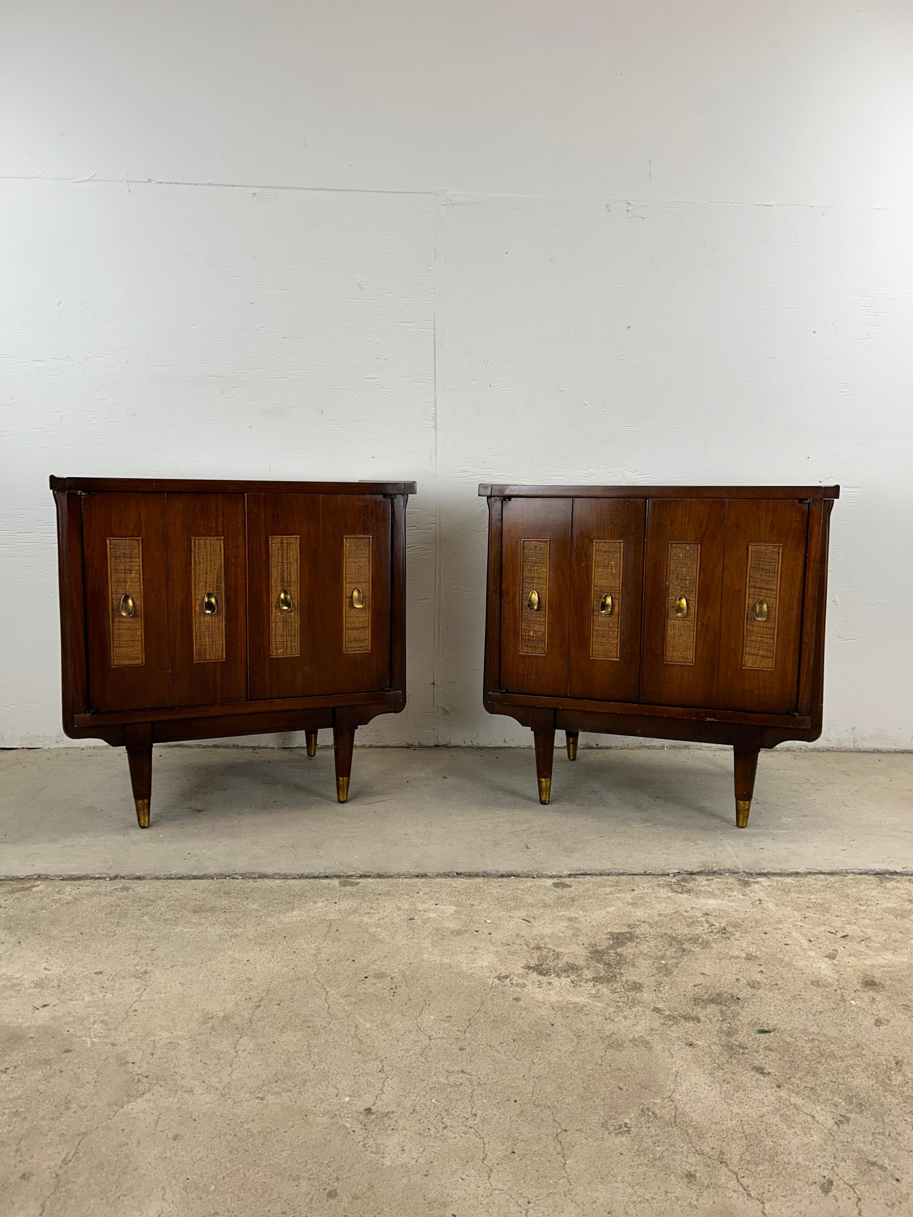 This mid century modern pair of nightstands features hardwood construction, walnut veneer with original finish, two cabinet doors with caned accent and brass hardware, interior shelving, atop brass capped tapered legs.   Matching lowboy dresser with