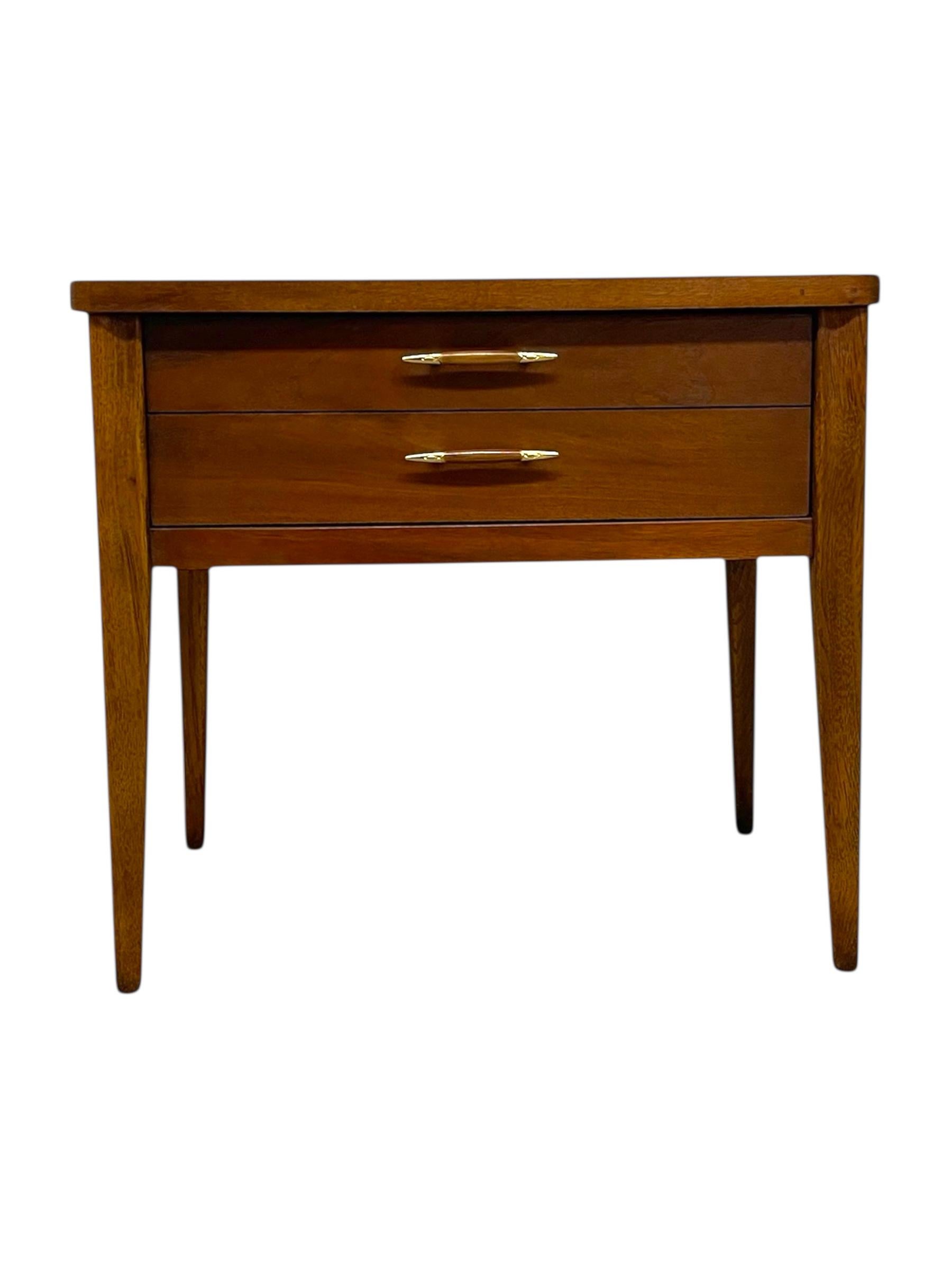 This vintage Mid-Century Modern end table features clean lines, tapered legs, and a warm wood finish characteristic of 1960s American design. The piece offers  smoothly operating drawer, each fitted with brass-tone handles that add a subtle metallic