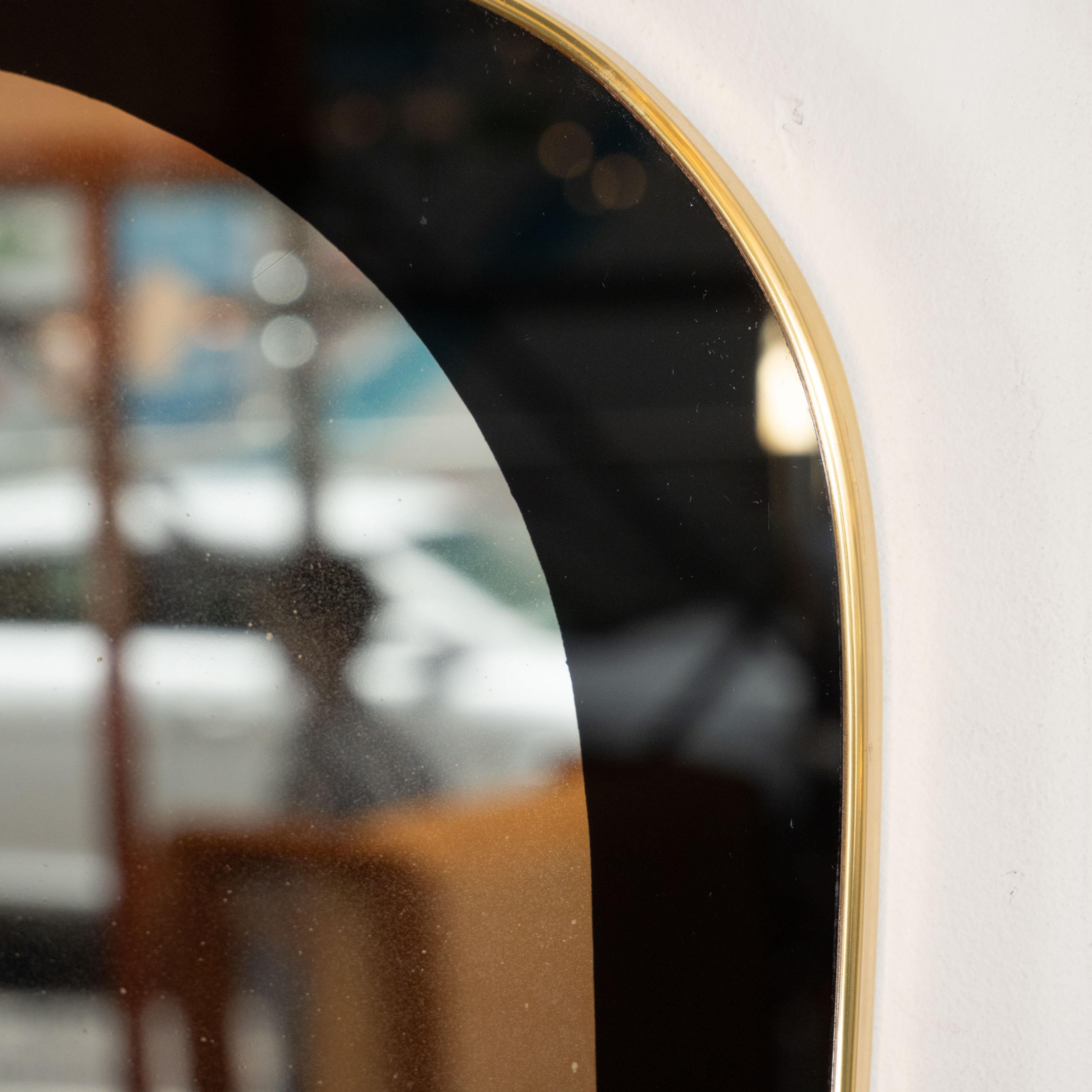 This stunning and sophisticated wall mirror was realized in Italy, circa 1970. It features an atomic oval form wrapped in brass. The center of the mirror is clear while the borders have been reverse églomisé painted in a rich opaque onyx hue. With