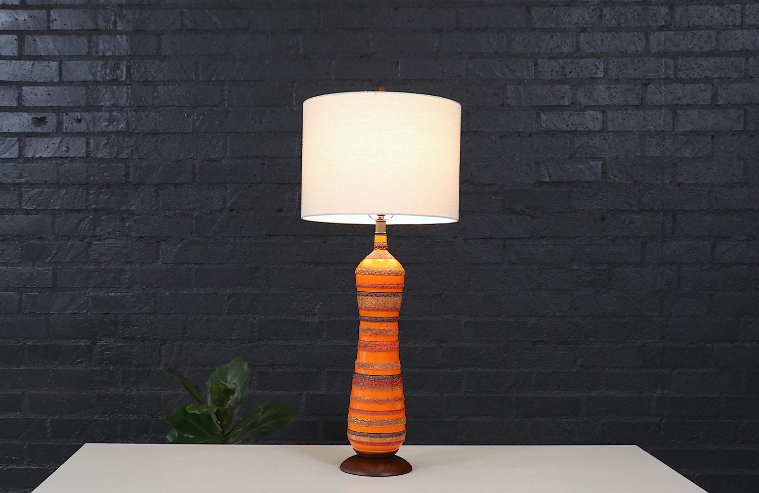 Esta llamativa lámpara de sobremesa Mid-Century Modern de Phil-Mar es una mezcla perfecta de diseño atrevido y artesanía experta. El cuerpo de cerámica presenta un vibrante esmalte naranja con bandas texturizadas similares a la lava, que añaden