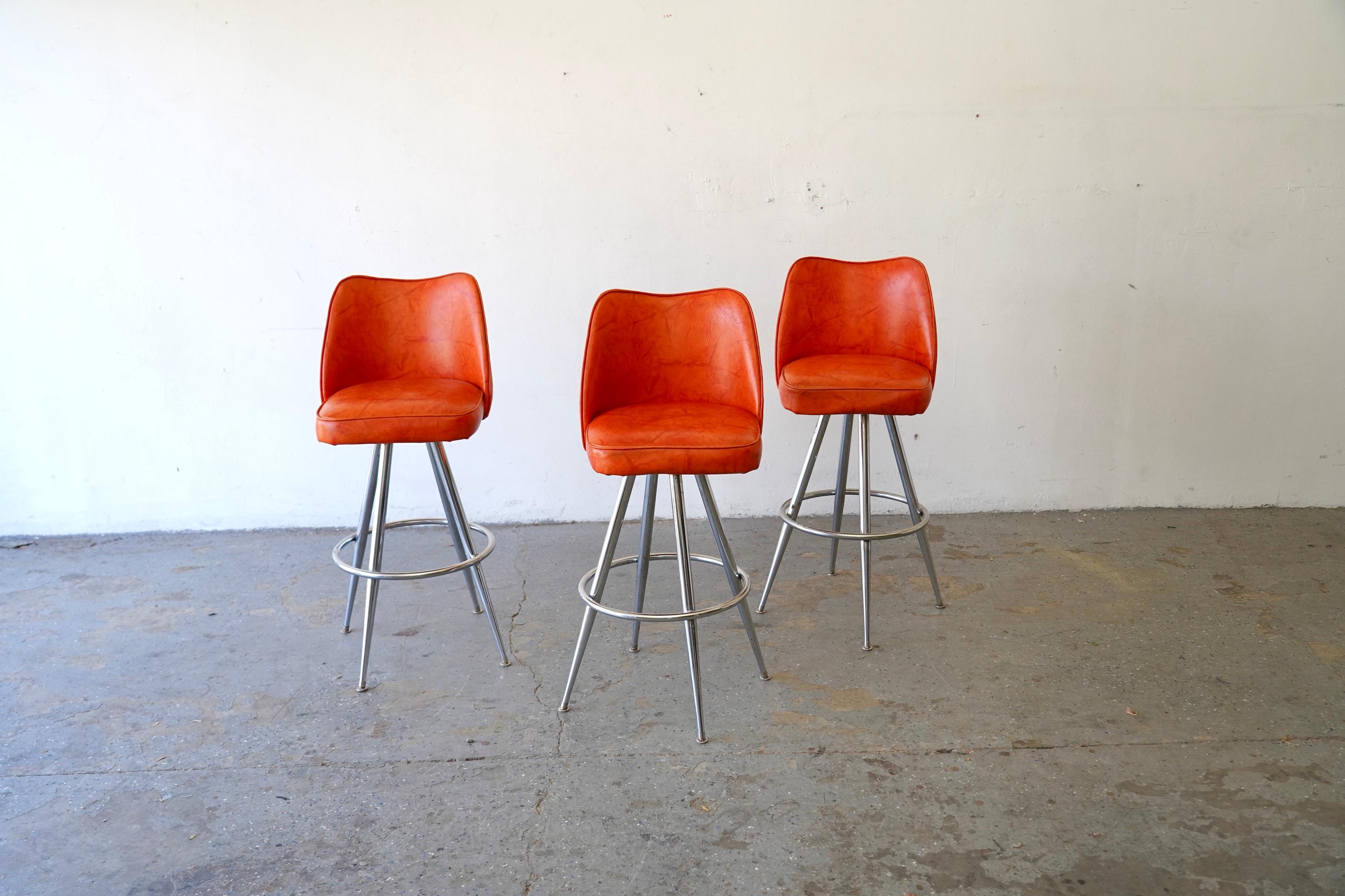 Striking set of three Mid-Century Modern bar stools, featuring bold orange vinyl upholstery and sleek chrome-plated bases. The curved low-back design offers both style and comfort, with the original vinyl exhibiting rich vintage character and subtle