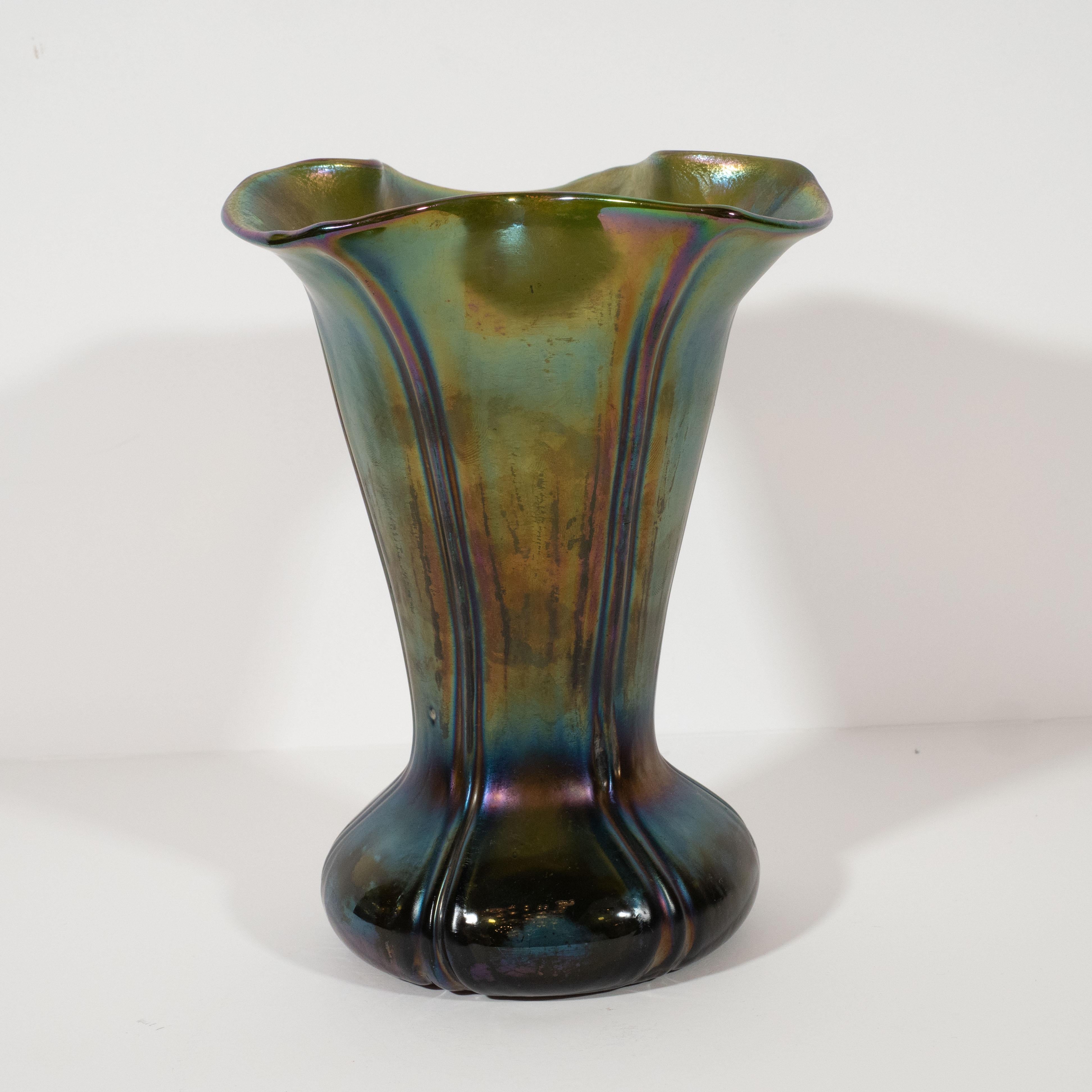 This stunning handblown favrile glass vase was realized in the United States, circa 1970. It features a reeded hourglass form, cinched at its waist, with a sinuously curved mouth. The favrile glass exhibits a beautiful iridescent quality offering