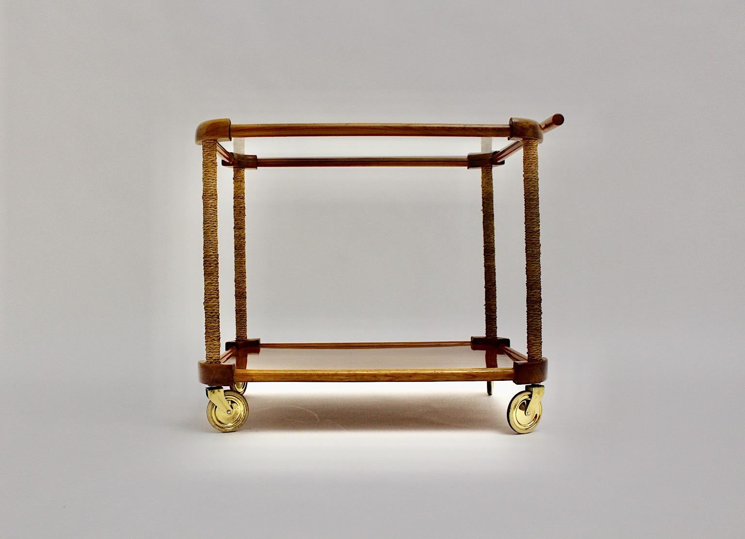 Mid-Century Modern organic vintage bar cart or tea cart from cherry, brass and cords by Max Kment Kunstgewerbliche Werkstätten 1950s Austria.
While the bar cart shows two tiers, one tier made from clear glass and one tier from plywood. Wrapped