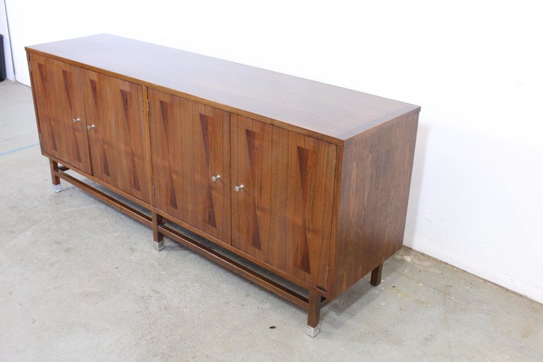 Offered is a Mid-Century Modern walnut credenza by Paul Browning for Stanley Furniture. It features 2 shelves and 3 drawers. It has an hourglass inlay on the four doors. The right side which has 3 drawers has a felted storage area. In very good