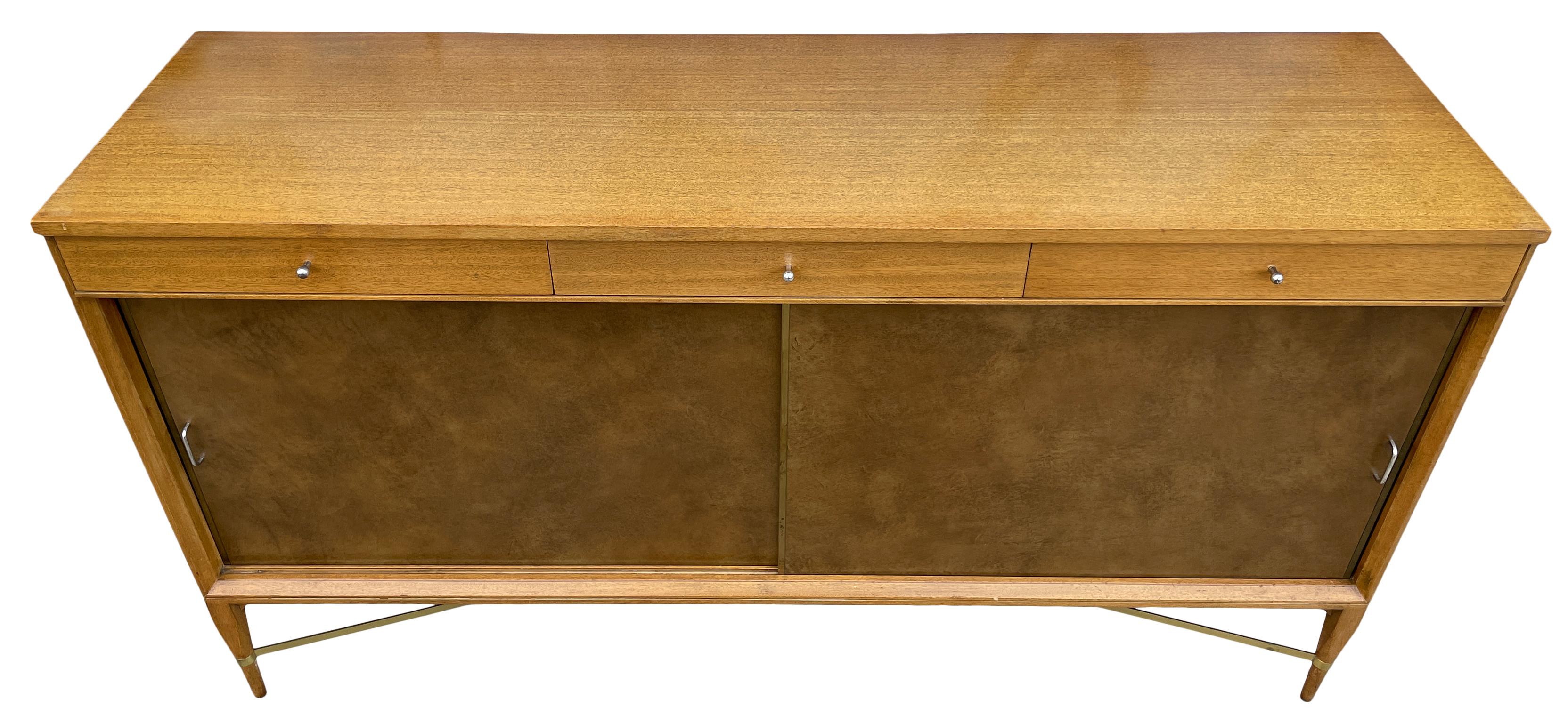 An exceptional Mid-Century Modern brown leather sliding doors sideboard/credenza with 3 top drawers. Behind the sliding doors are 2 adjustable shelves in a dark brown finish. The outside of the credenza is a lighter blonde finish. Designed By Paul