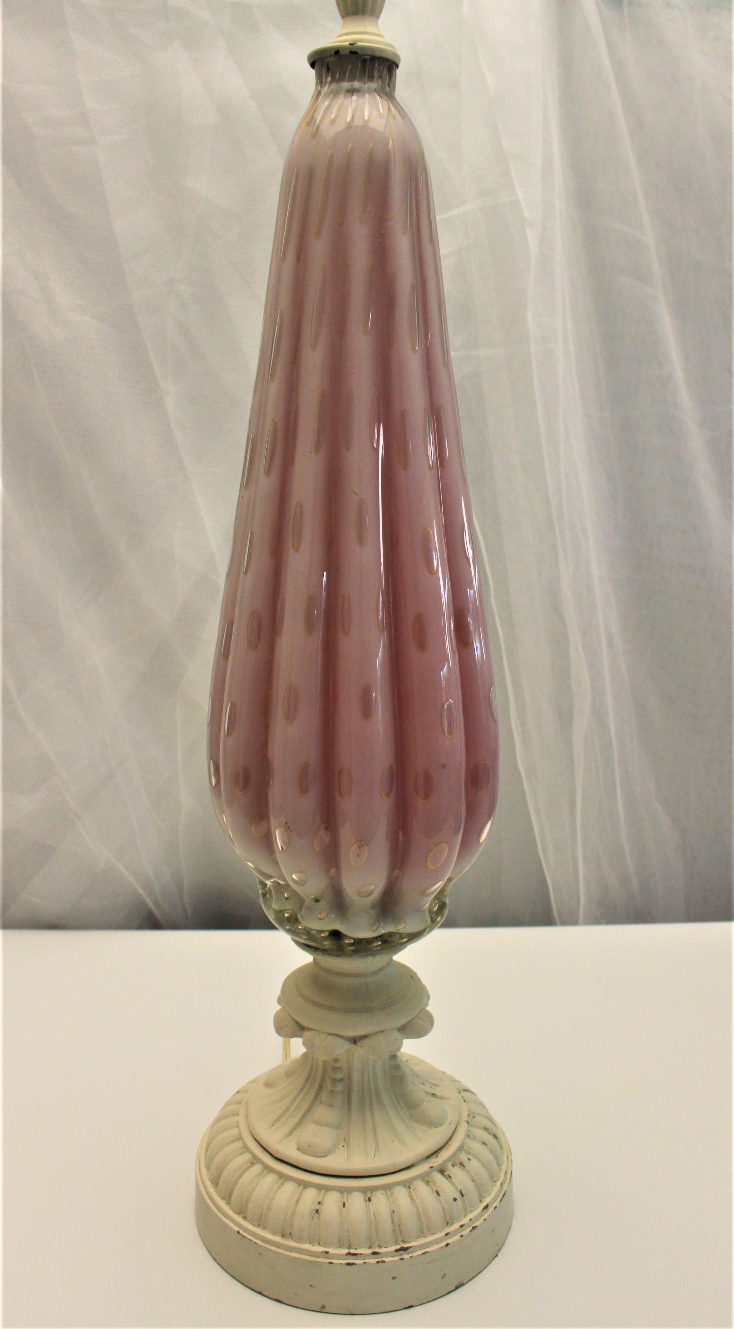 This Mid-Century Modern art glass table lamp is done with pink art glass with controlled bubbles and gold flecks or aventurine with a tapered and ribbed body and accented with an off white painted metal top and base. It is presumed this lamp was