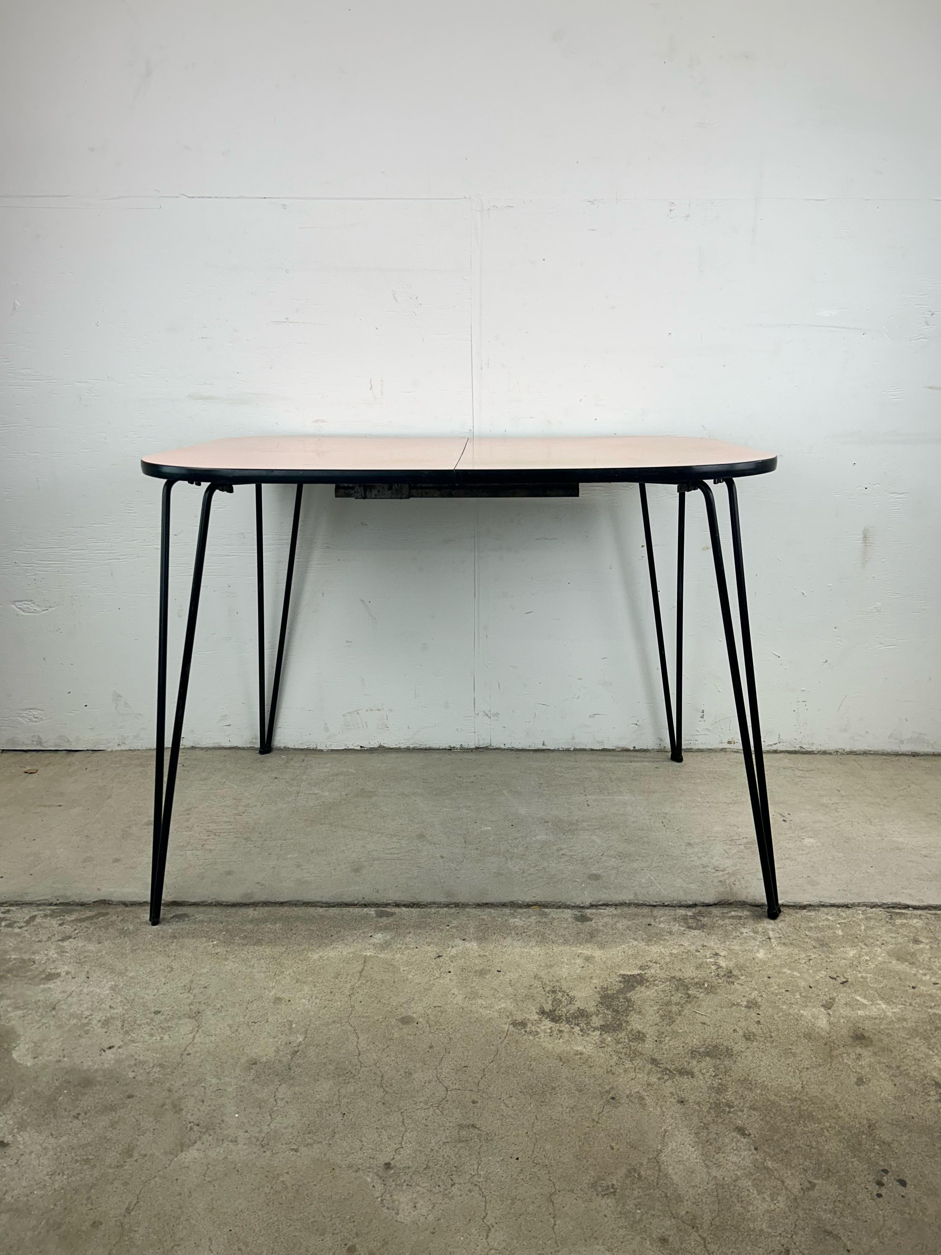 This mid century modern kitchenette table features formica construction, pink and white finish with some fading on the surface, black metal hairpin legs and one leaf.  This is your quintessential 1950s diner table.

Table with leaf measures 47.25w. 