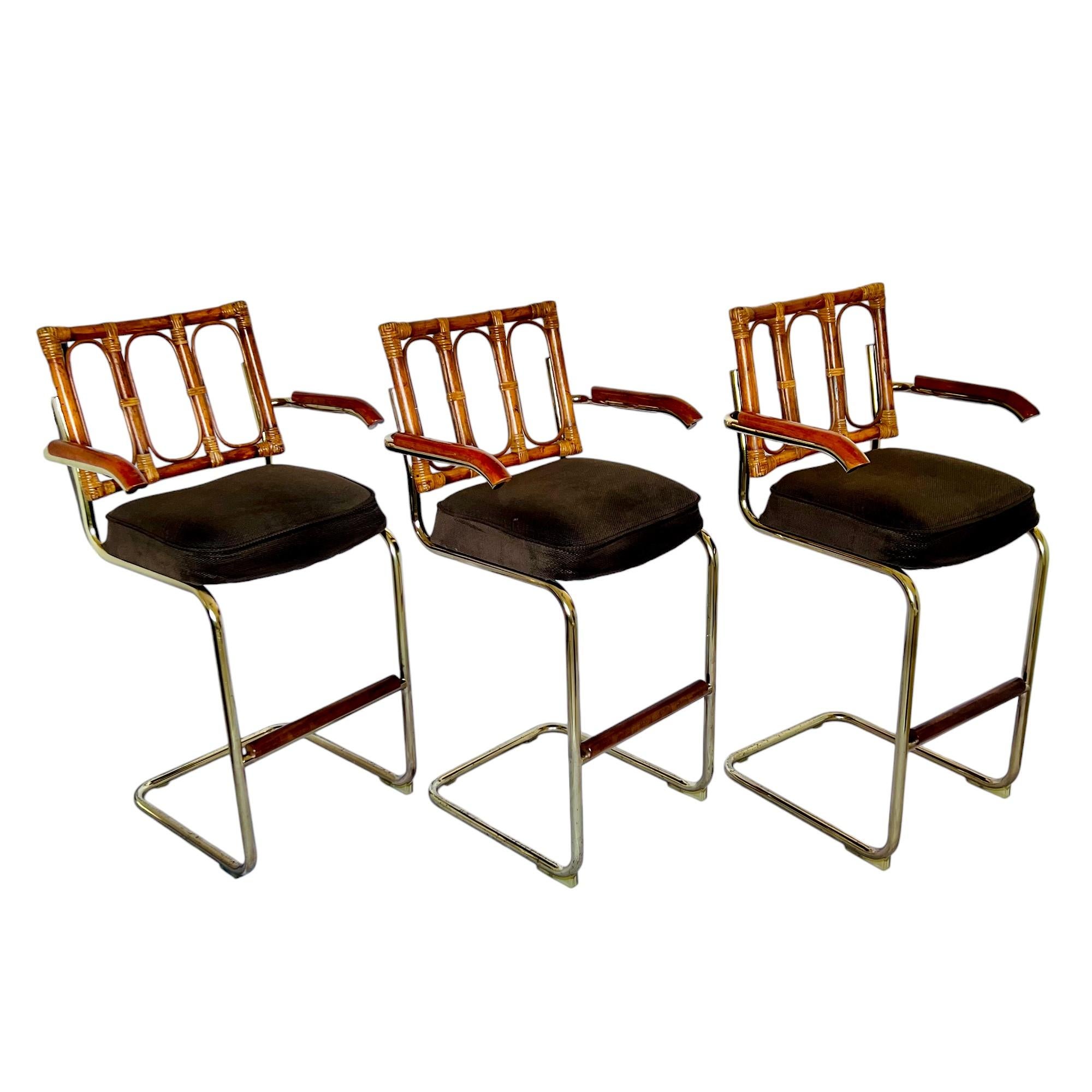 A vintage set of three Marcel Breuer Cesca style rattan and brass bar stools with arms. The mid-century modern design features a tubular brass finish metal frame, rattan back with looped and wrapped details, a thick cushioned seat in a dark brown