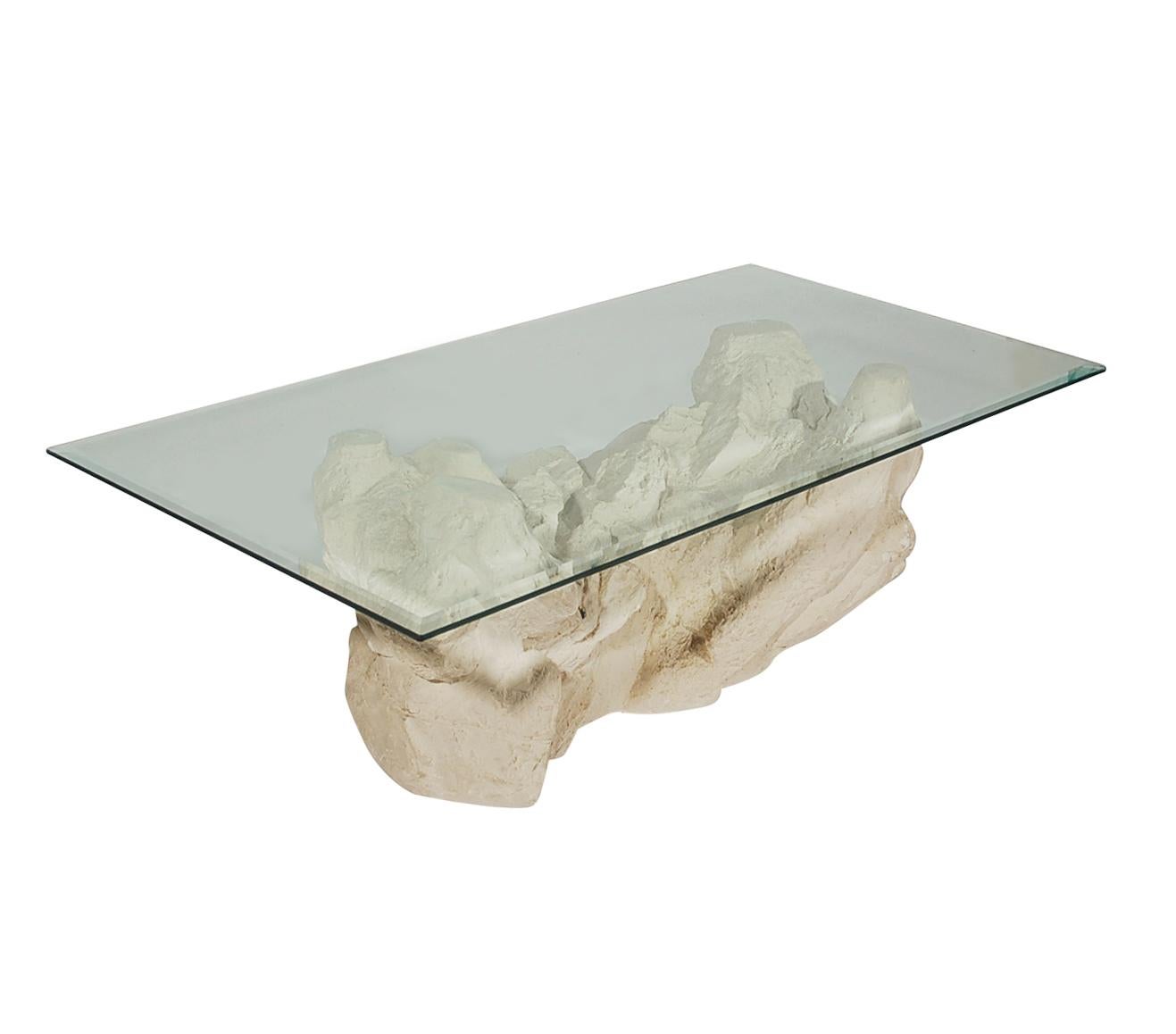 A sculptural faux rock formation table made by Sirmos in the 1970s. It features a plaster molded rock formation base with a clear glass top.