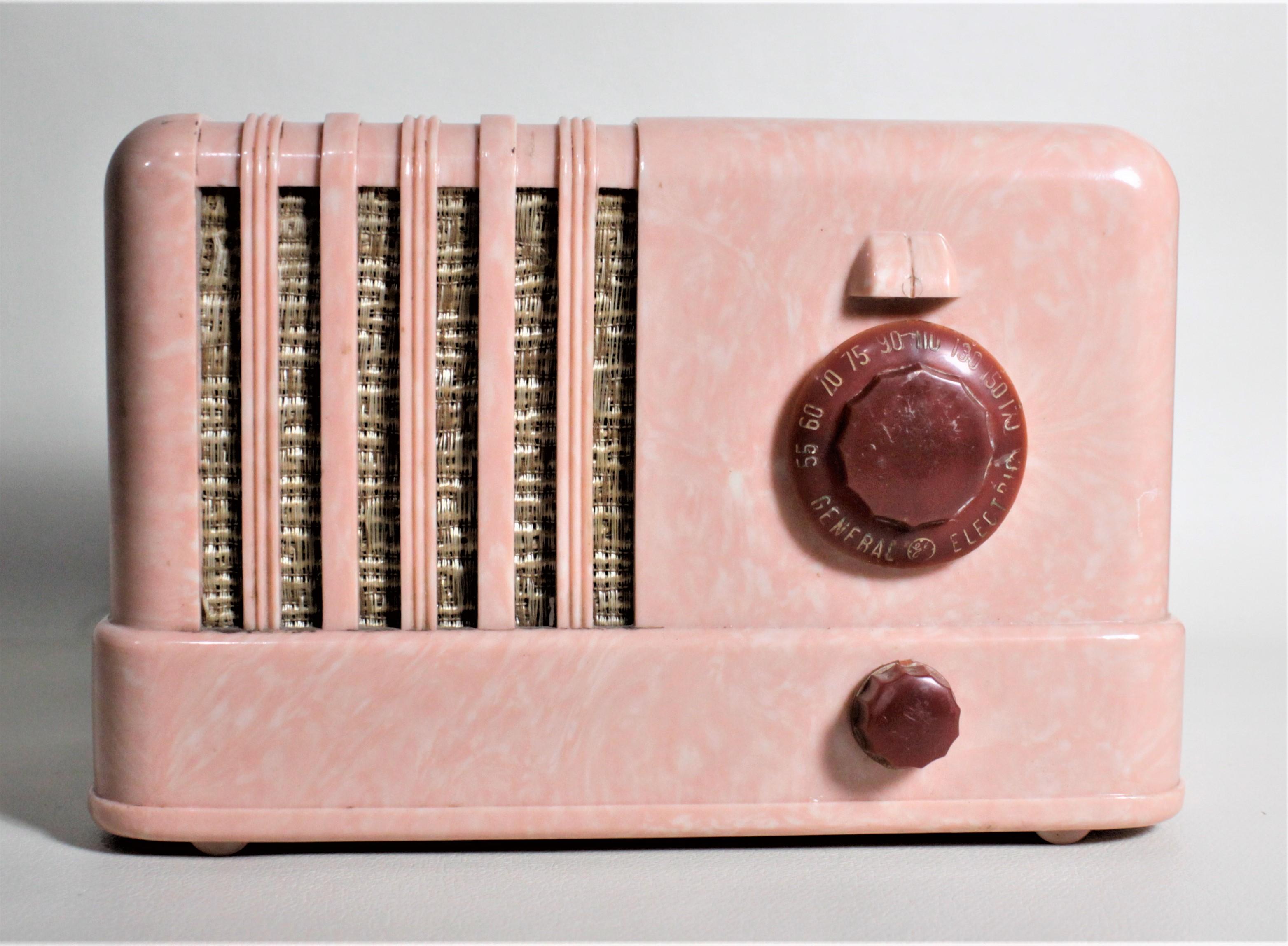 This table radio was produced by the Canadian General Electric Company in approximately 1960 in the period Mid-Century Modern style. The case of the radio is done in a bright marbled bubble gum pink and white, with contrasting blood red or burgundy