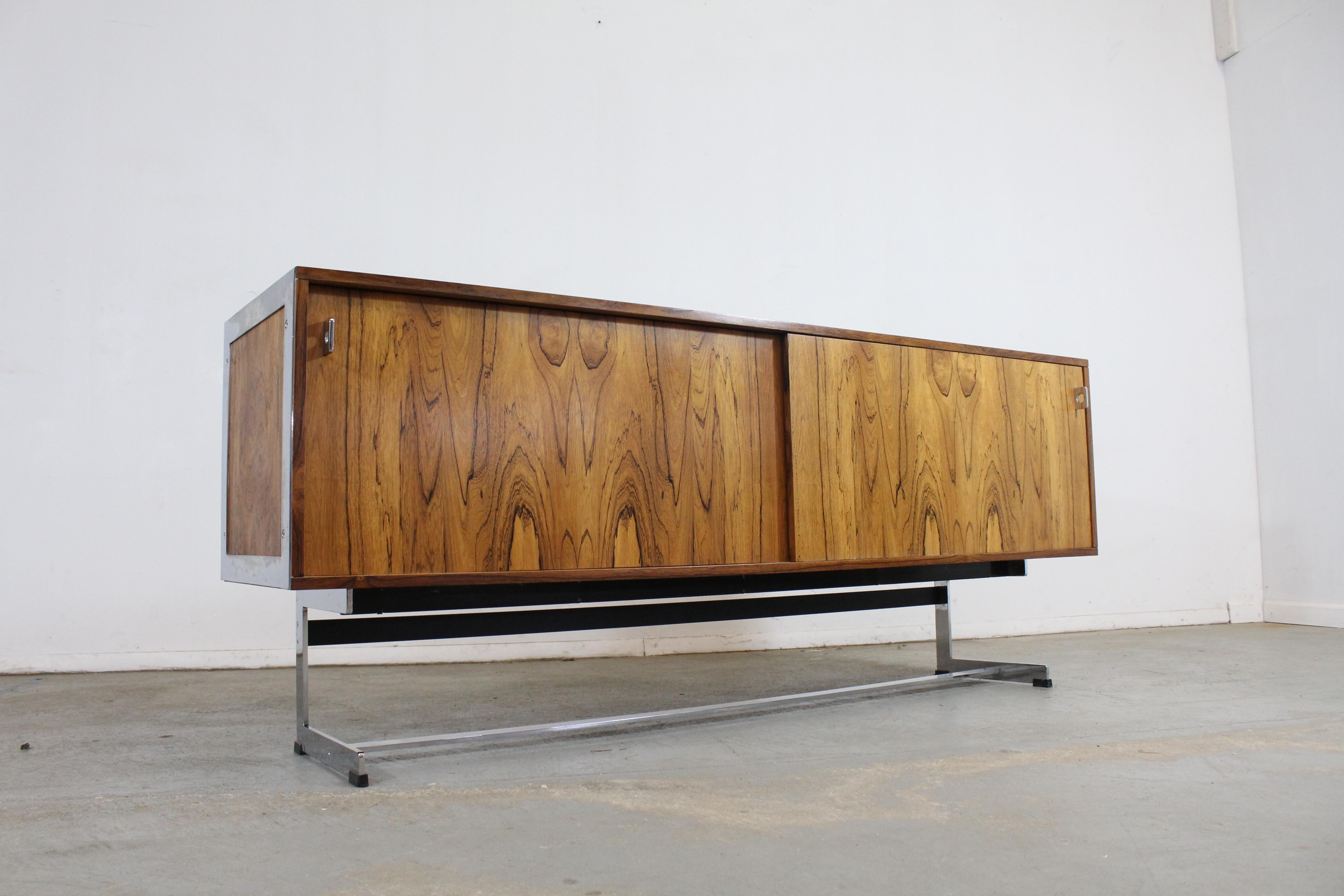 Offered is a beautiful Mid-Century Modern credenza, designed by Richard Young for Merrow Associates. It has chrome legs and pulls. Features two sliding doors with glass shelving on both sides. It is made of full grain wood. It's in very good