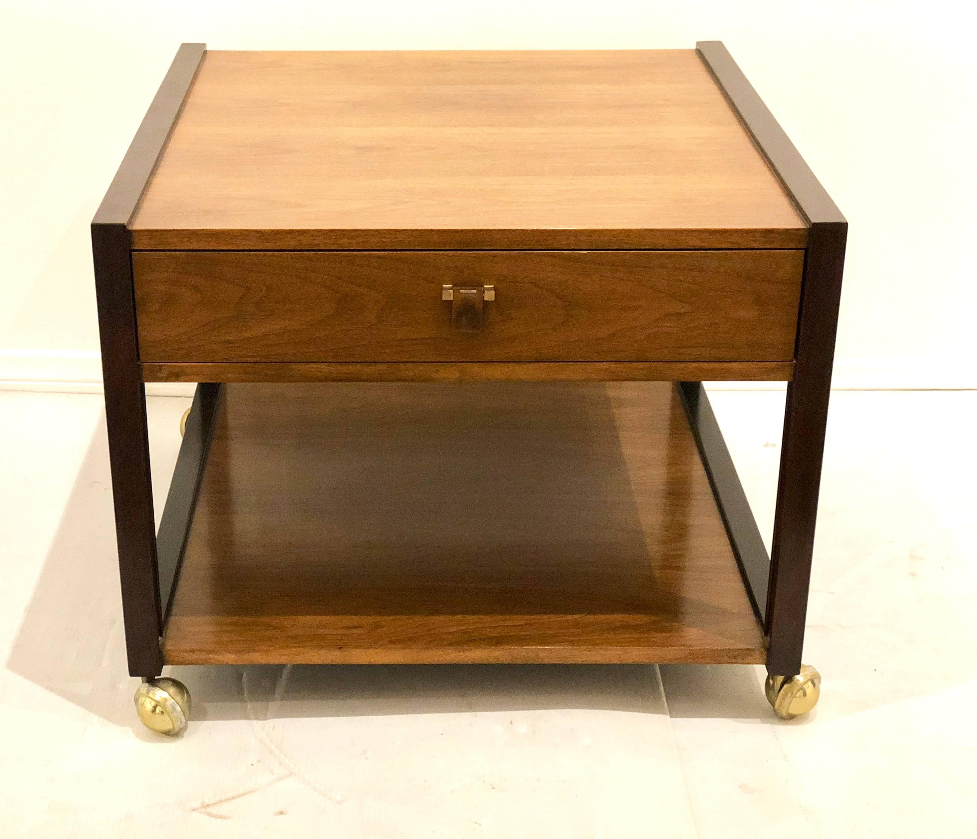 The design of this wide side table by Edward Wormley for Dunbar, in a walnut and rosewood combo, drawer with a rosewood and brass pull, a deep shelf below and bold brass ball casters for easy moving. Great in between two club chairs.