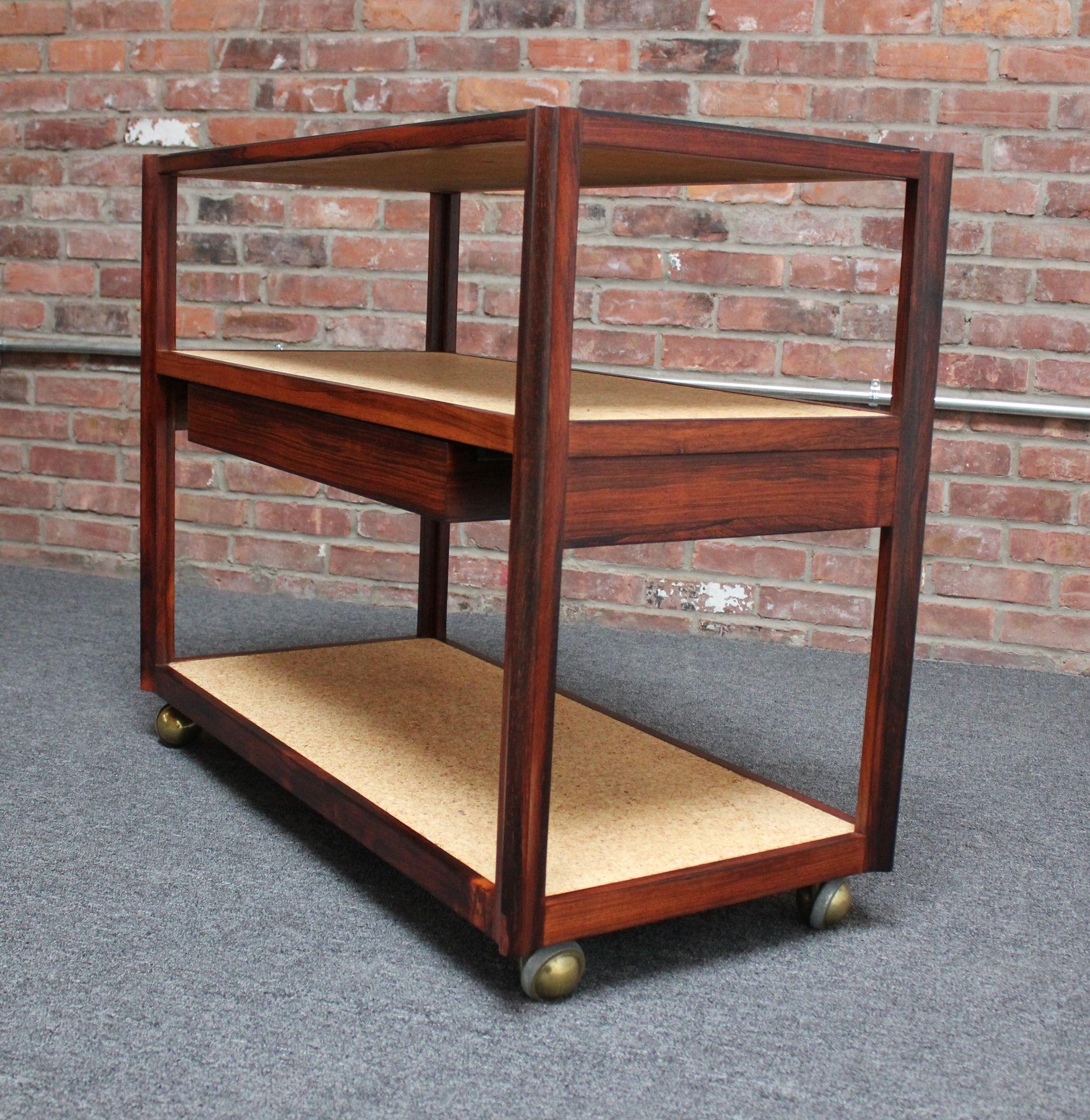 Three-tiered bar cart/tea trolley Model No. 6409 designed by Roger Sprunger for Dunbar (Indiana, USA, circa 1960s).
Composed of a inset slate top with two lower cork surfaces and a shallow drawer for accessories, all supported by brass caster wheels