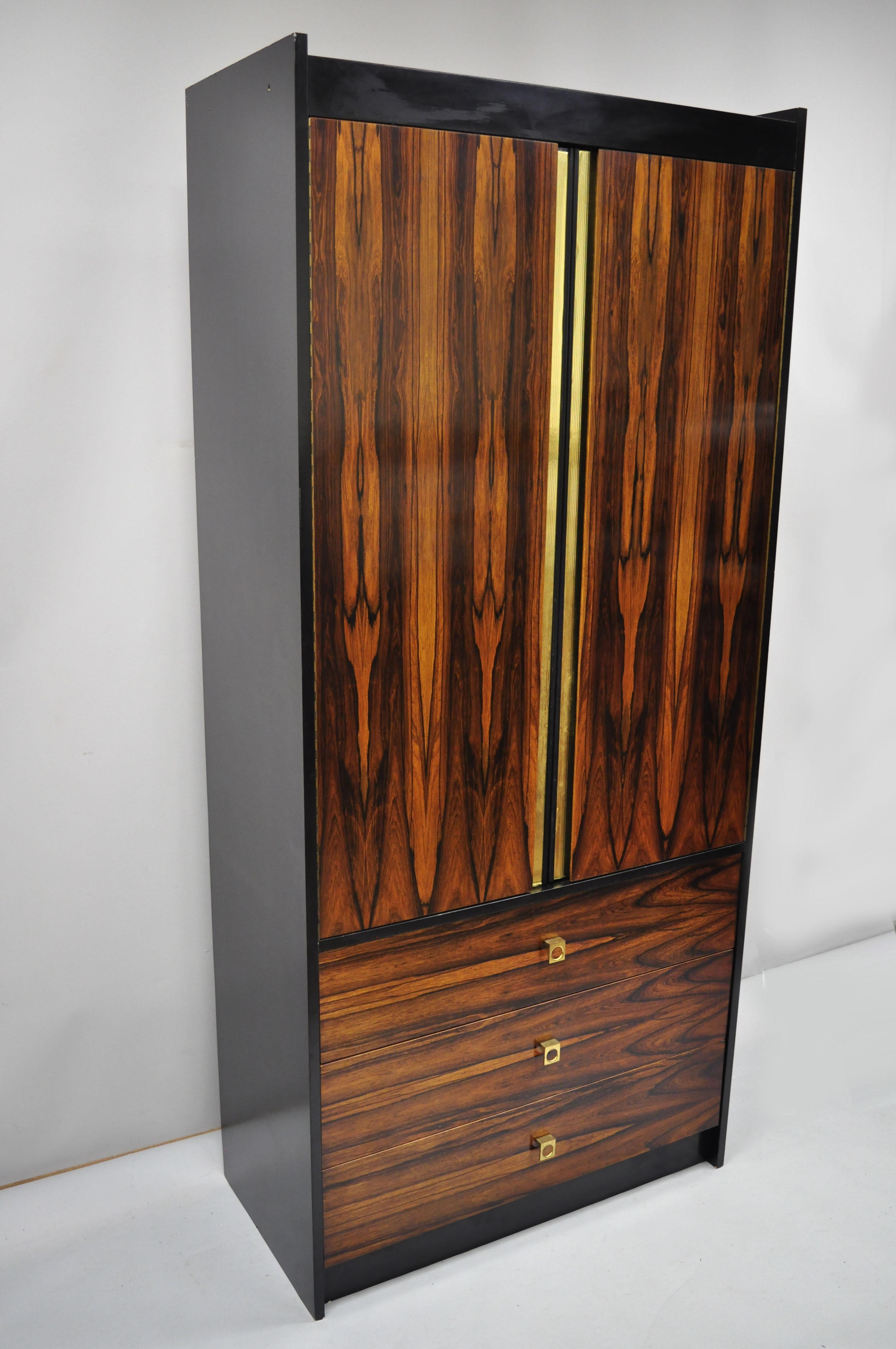 Vintage Mid-Century Modern rosewood and black laminate John Stuart style wardrobe cabinet armoire (B). Item features a tall impressive form, stunning wood grain, 2 adjustable shelves, 1 fixed shelf, gold foil taped trim, 2 swing doors, 3 drawers,