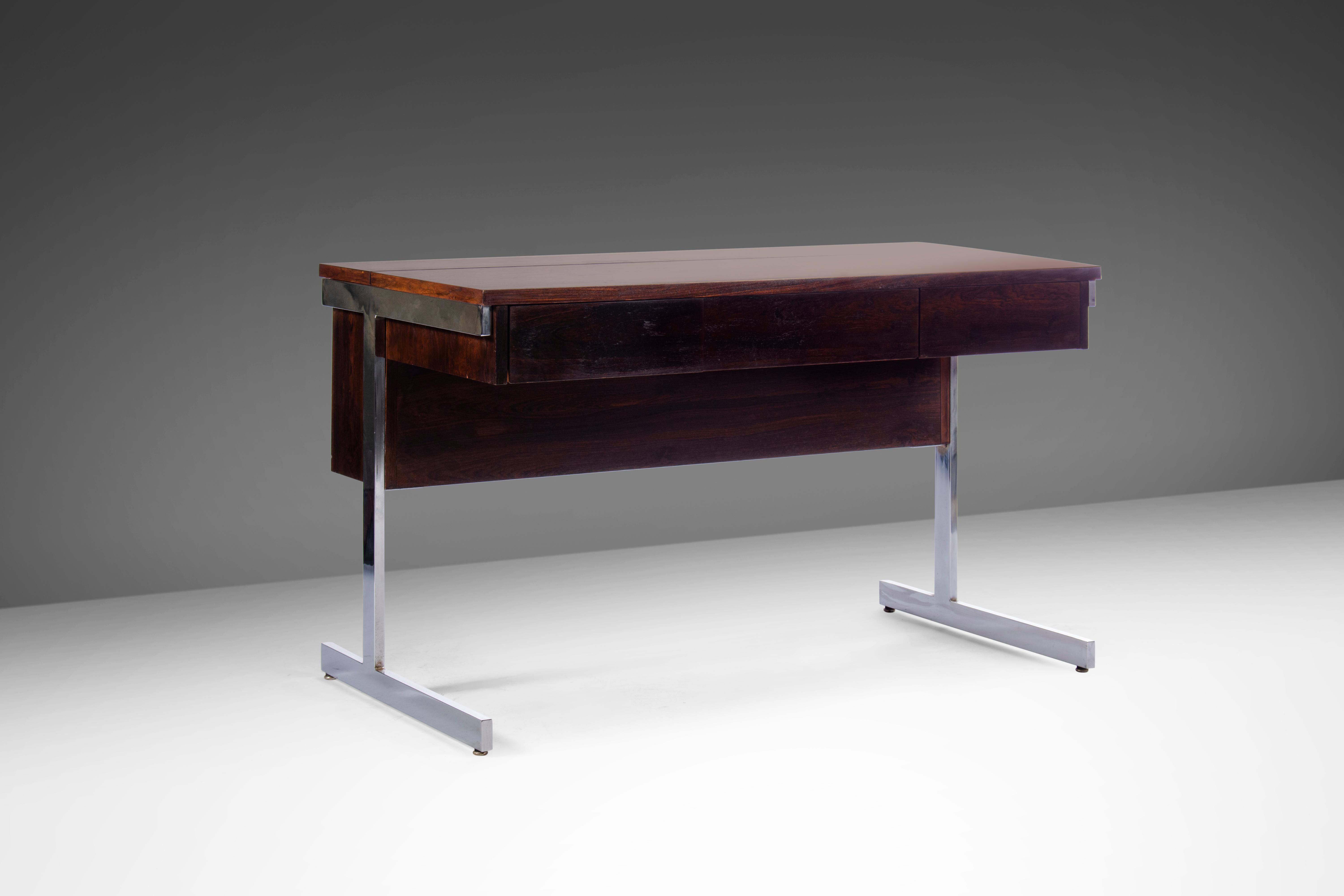Constructed from rosewood and set on a T-shaped chrome base. There are three front flat faced drawers and a flush lift-top hidden compartment at the back that can be used for files or storage. Circa 1970's.
   
