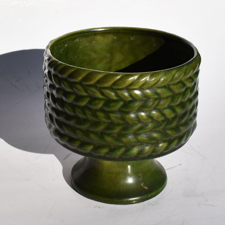 A round textural green planter in green. Round in form, this lovely jardinière stands on a small round base. The exterior of the planter is decorated with a braided pattern around the sides. The entire piece is glazed in a matte green color.

We