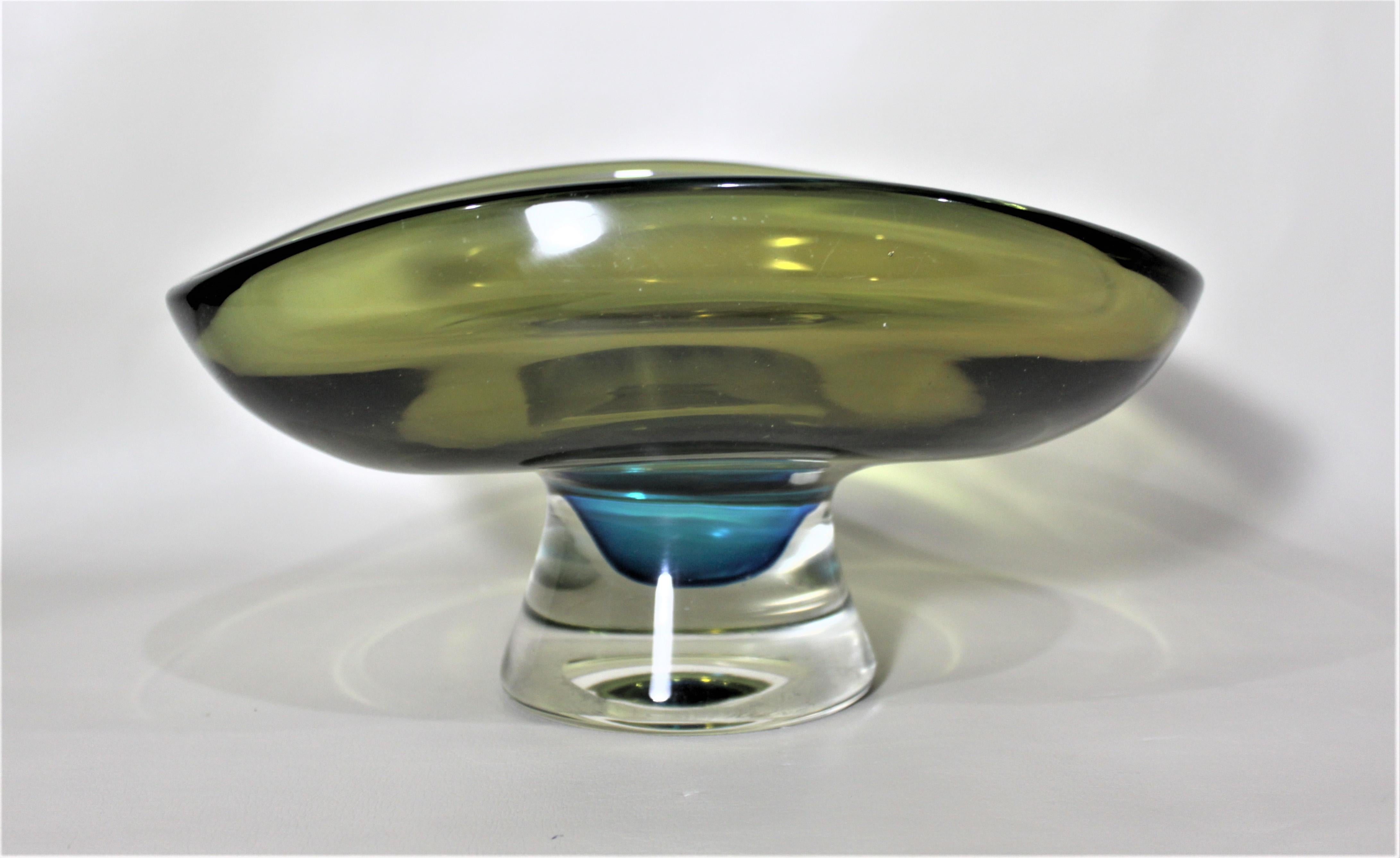 This Mid-Century Modern art glass pedestal, or centerpiece bowl is unsigned but presumed to have been made by Kosta Boda. The bowl is done in a deep olive green with a clear pedestal base with a cobalt blue inclusion.