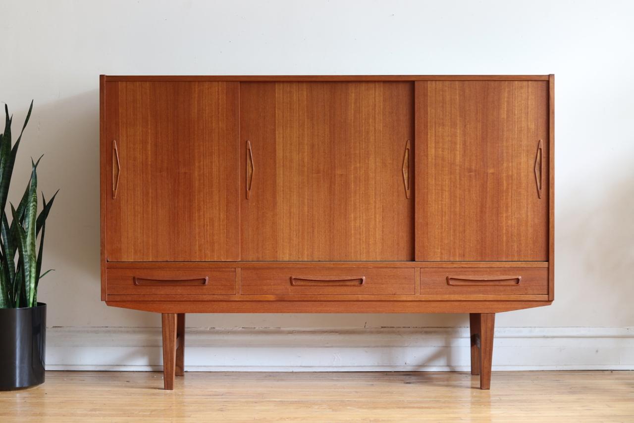 Mid-Century Modern Danish Modern tall teak wood credenza.
Just imported from Copenhagen!
Sliding cabinet doors hold adjustable shelving. 
Three dovetailed drawers.
Triangle/arrow shaped handles.
Excellent vintage condition.

Measures: 64 3/4”