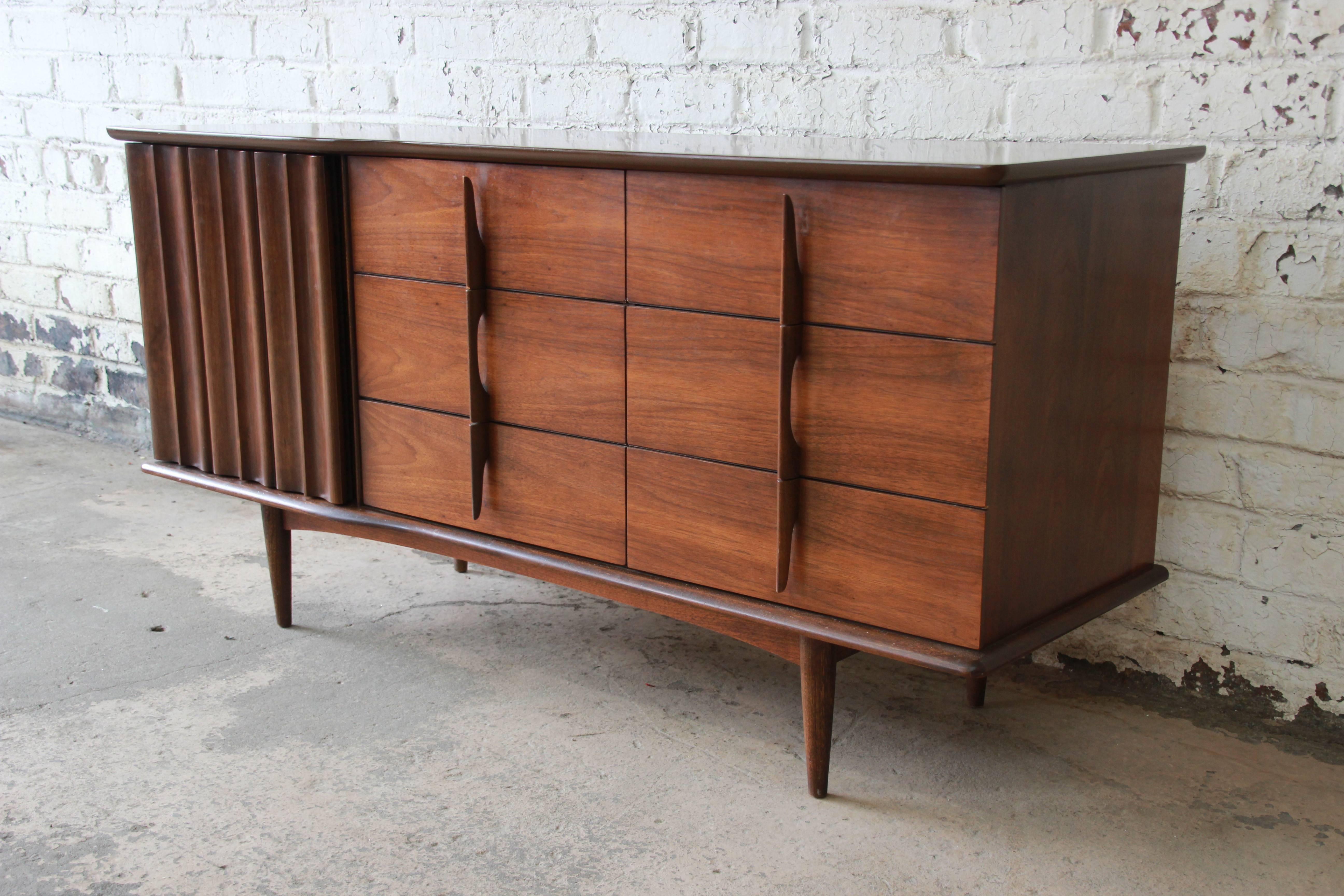 A stylish Mid-Century Modern sculpted walnut triple dresser or credenza by United Furniture Co. The dresser features gorgeous walnut wood grain and sleek Mid-Century Modern design. It offers ample room for storage, with nine dovetailed drawers. The