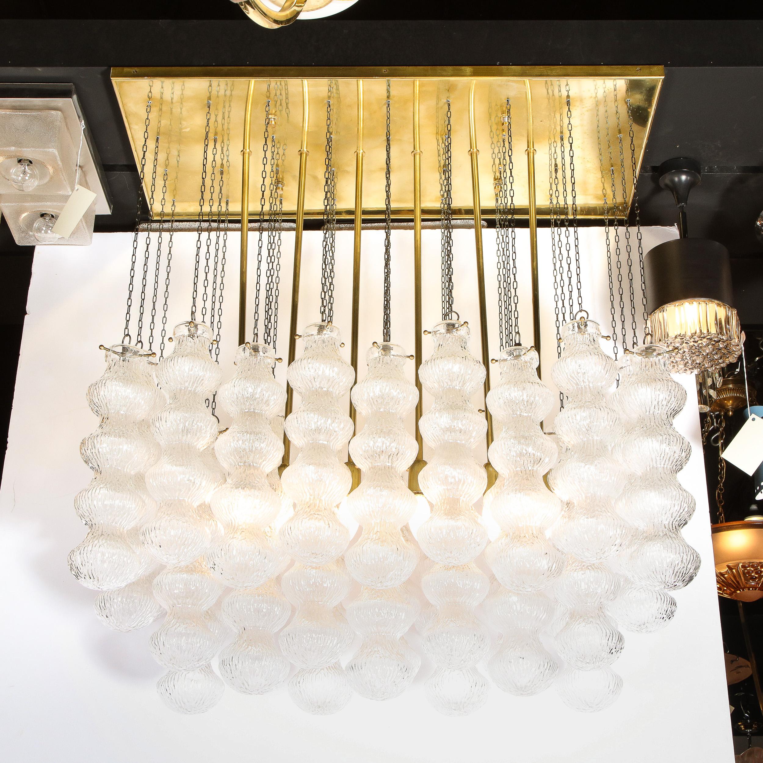 This stunning Mid-Century Modern chandelier was realized in Murano, Italy- the island off the coast of Venice renowned for centuries for its superlative glass production. It features a rectangular polished brass backplate with six cylindrical rods