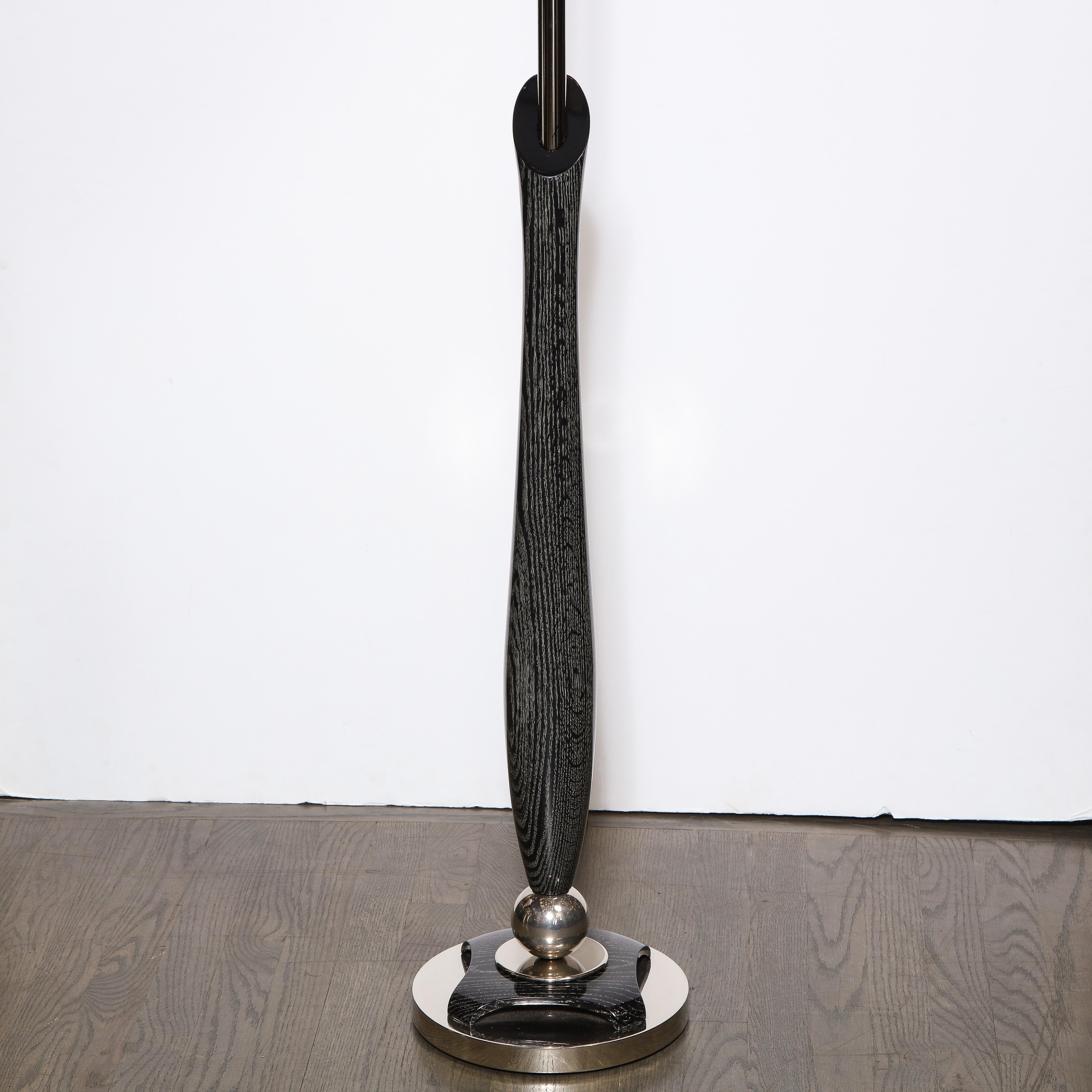 This striking Mid-Century Modern floor lamp was realized in the United States circa 1950. It features a inverse hourglass form silver cerused body that flares at its center with an angled (top cut on a bias) and brushed aluminum rod piercing the