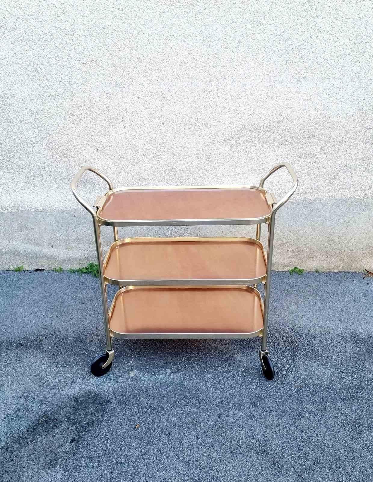 Ce très beau et élégant chariot de service a été fabriqué par Kaymet au Royaume-Uni dans les années 60.

Ce meuble est polyvalent et pratique. Il est composé d'un cadre en aluminium doré et de trois étagères/plateaux bruns. Le plateau supérieur, le