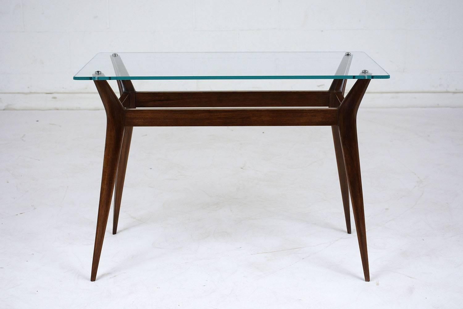 This 1960s Mid-Century Modern-style side table features a teak wood base stained in a rich walnut color with a lacquered finish. The table has a glass top with a flat polish attached to the stretched tapered legs. This side table is sturdy, sleek,