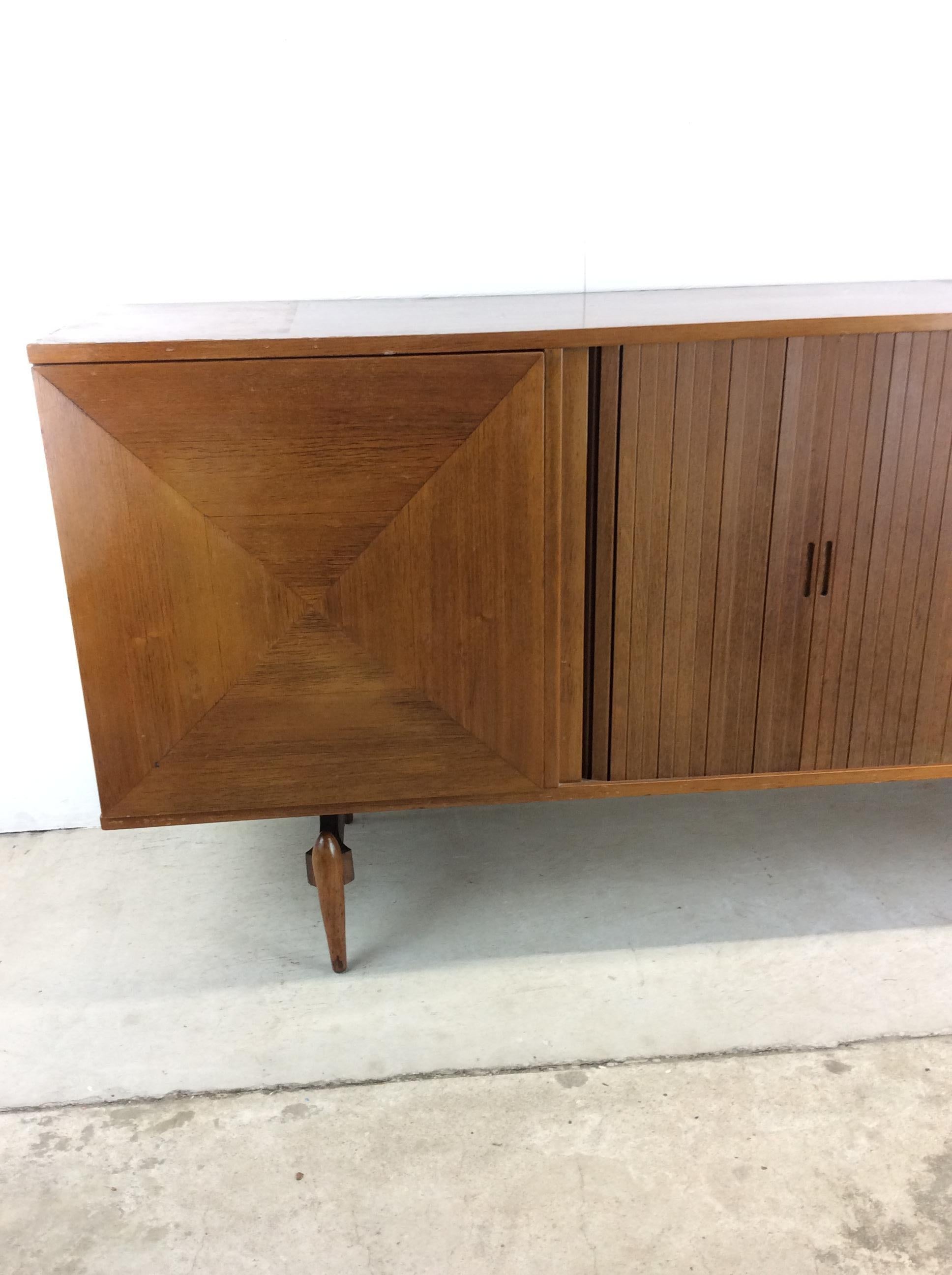 This mid century modern credenza features hardwood construction, walnut veneer with original finish, two diamond fronted cabinet doors with interior shelving, a tambour door with additional shelving, and a unique tapered legs.

Complimentary drop