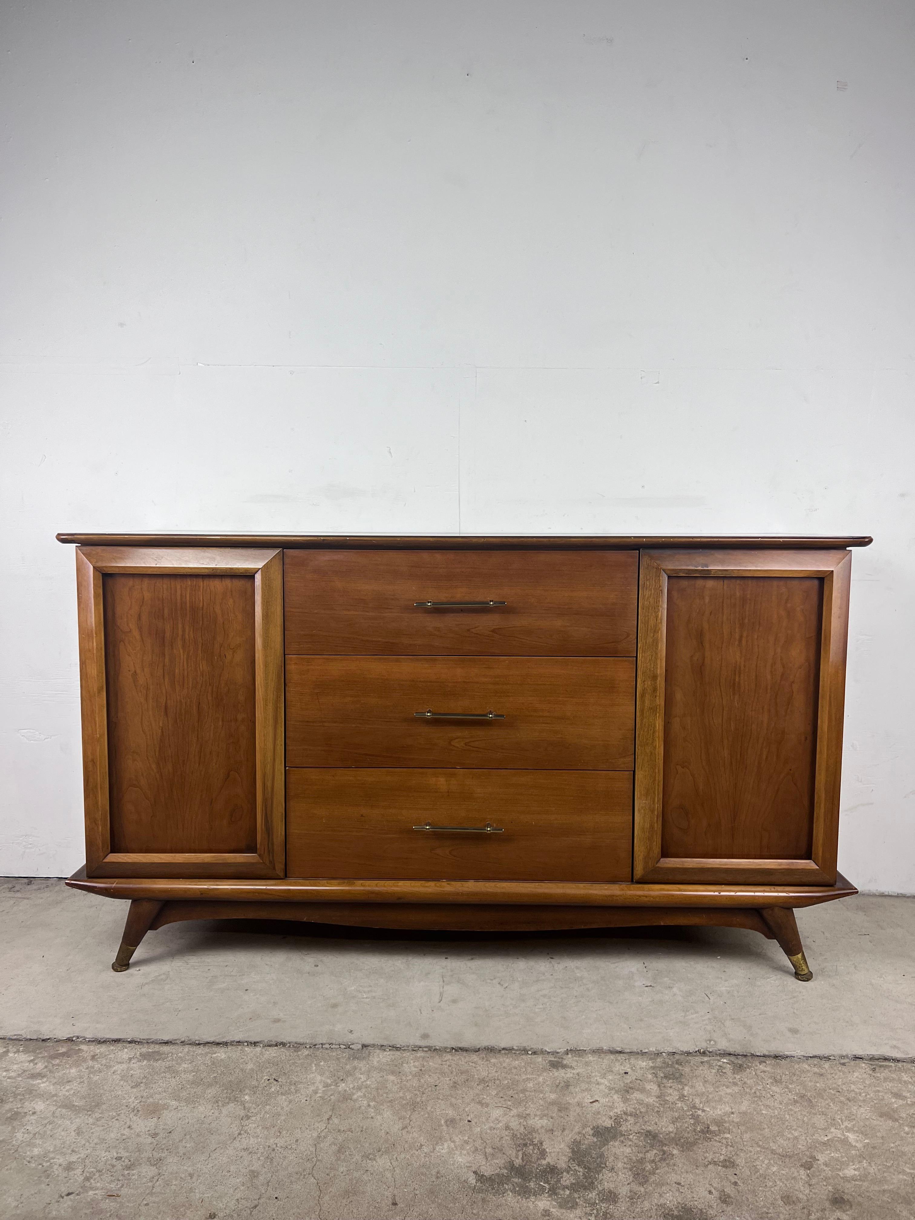 This mid century modern credenza features hardwood construction, walnut veneer with original finish, three dovetailed drawers with brass hardware, two cabinet doors with interior shelving, and brass capped tapered legs.