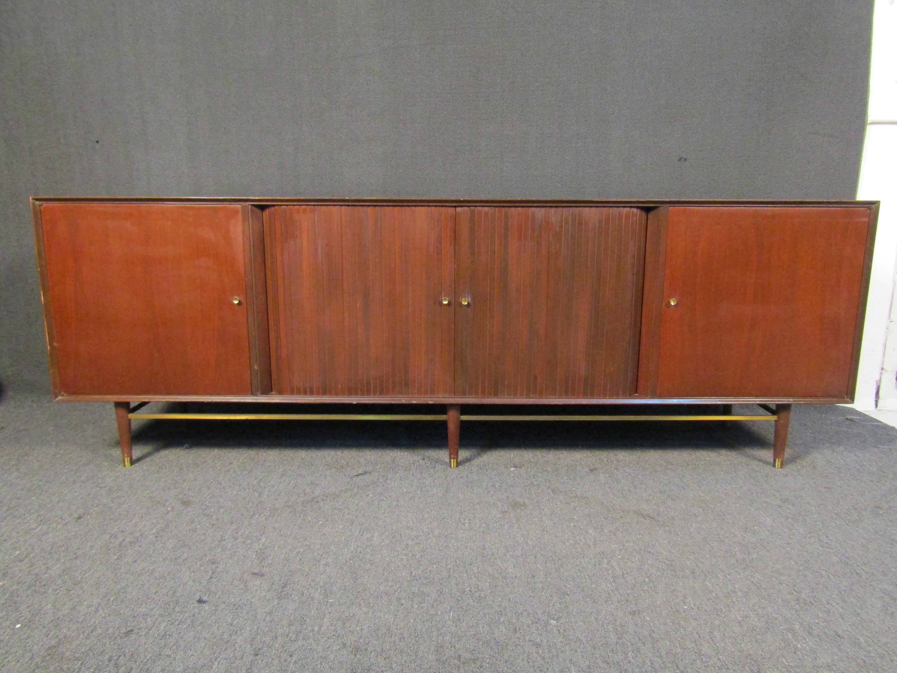 This large Mid-Century Modern combines rich woodgrain and quality materials with tambour doors that open to reveal plenty of storage. Brass accents and footing add an extra touch of vintage style. Please confirm item location with seller (NY/NJ).