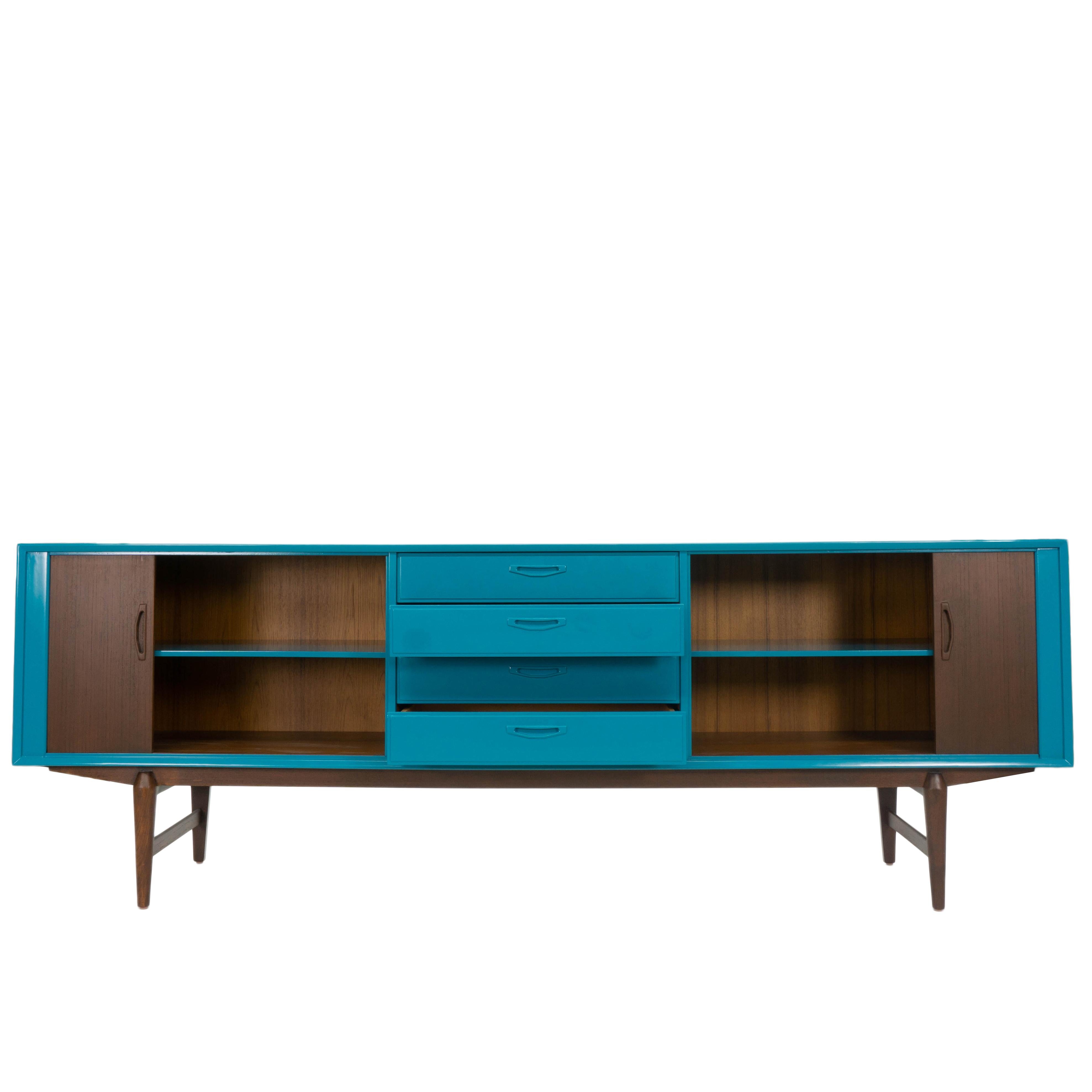 This vintage Mid-Century Modern sideboard has been refinished and lacquered in a bright turquoise blue. The wooden doors and tapered legs have a dark walnut stain with some red undertones. The sideboard also features two tambour doors and four
