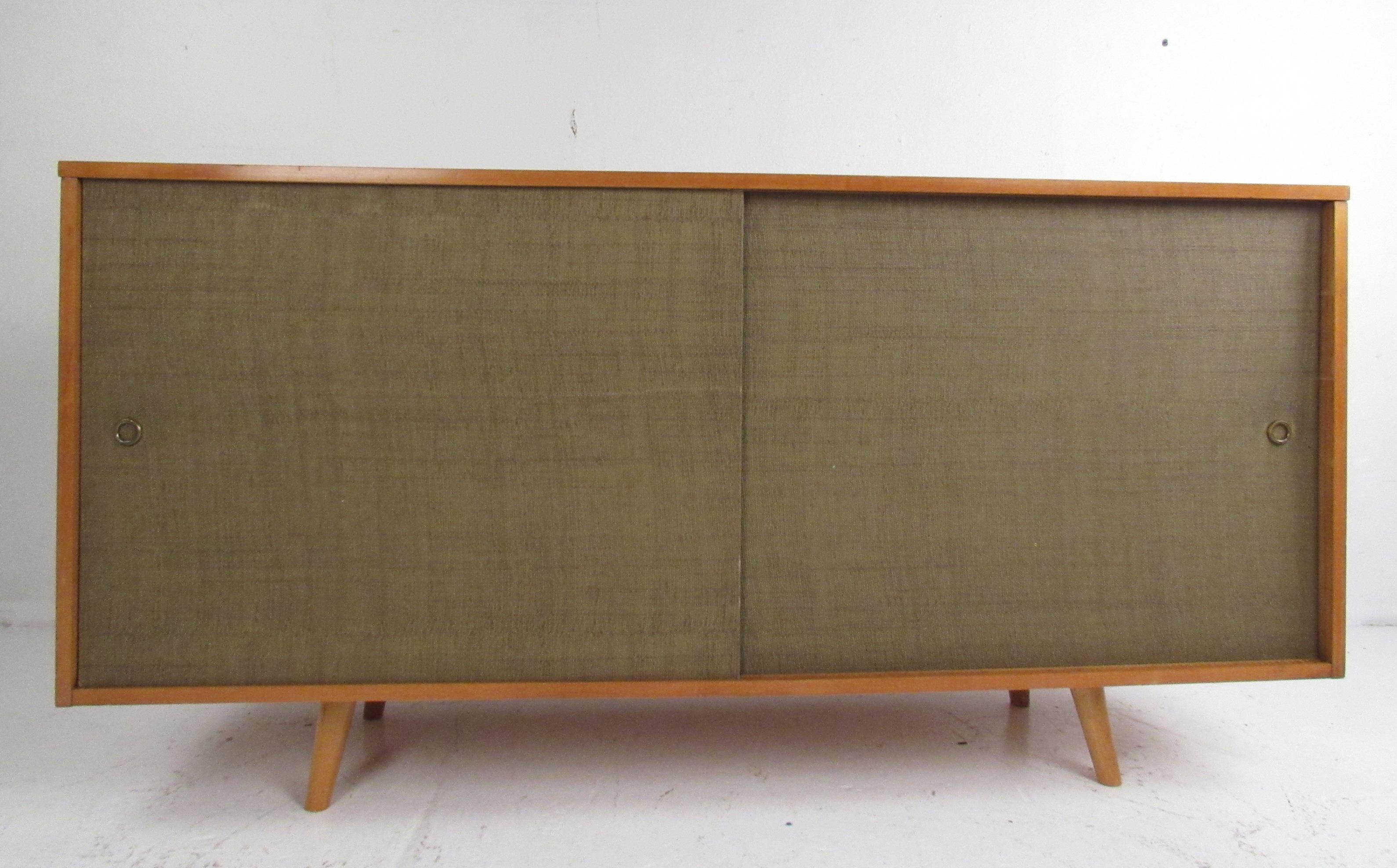 This stylish vintage modern sideboard boasts two sliding doors covered in grasscloth that open up to unveil ample storage space. A sleek design that boasts drawers with cut-out pulls, iconic splayed legs, and a vintage maple finish. This versatile