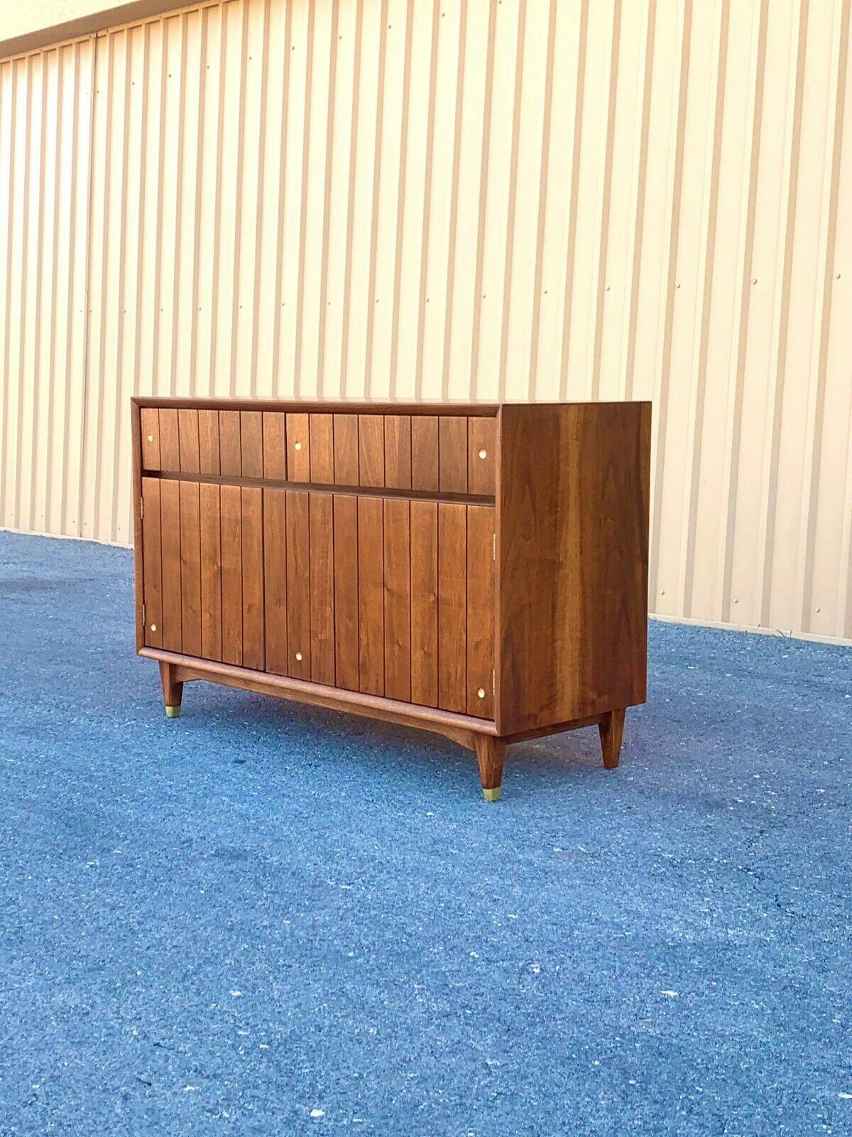 This vintage MCM credenza manufactured by Kroehler has a fixed panel in the center and two doors that open to reveal one continuous fixed shelf towards the back of the interior. Two shallow drawers lie above, one of which is compartmentalized for
