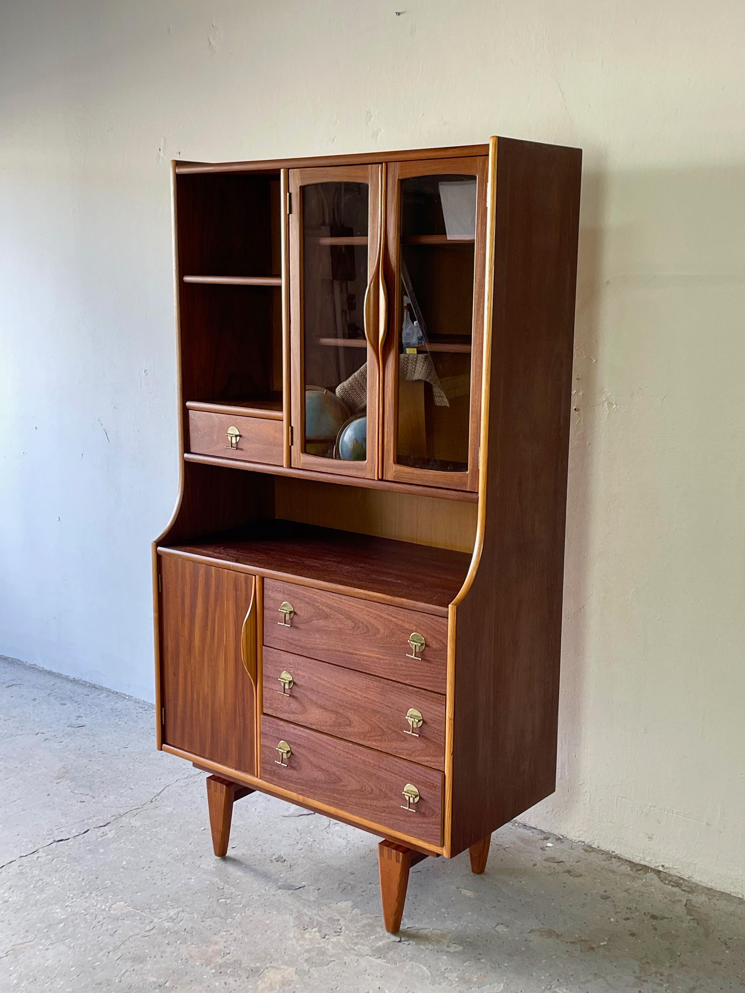 Mid-Century Modern walnut Stanley display hutch / china cabinet in a unique compact size. . This piece offers many unique features dovetailed tapered wood legs, two tone wood, glass doors with unique curved handles and brass pulls. Lower cabinet has