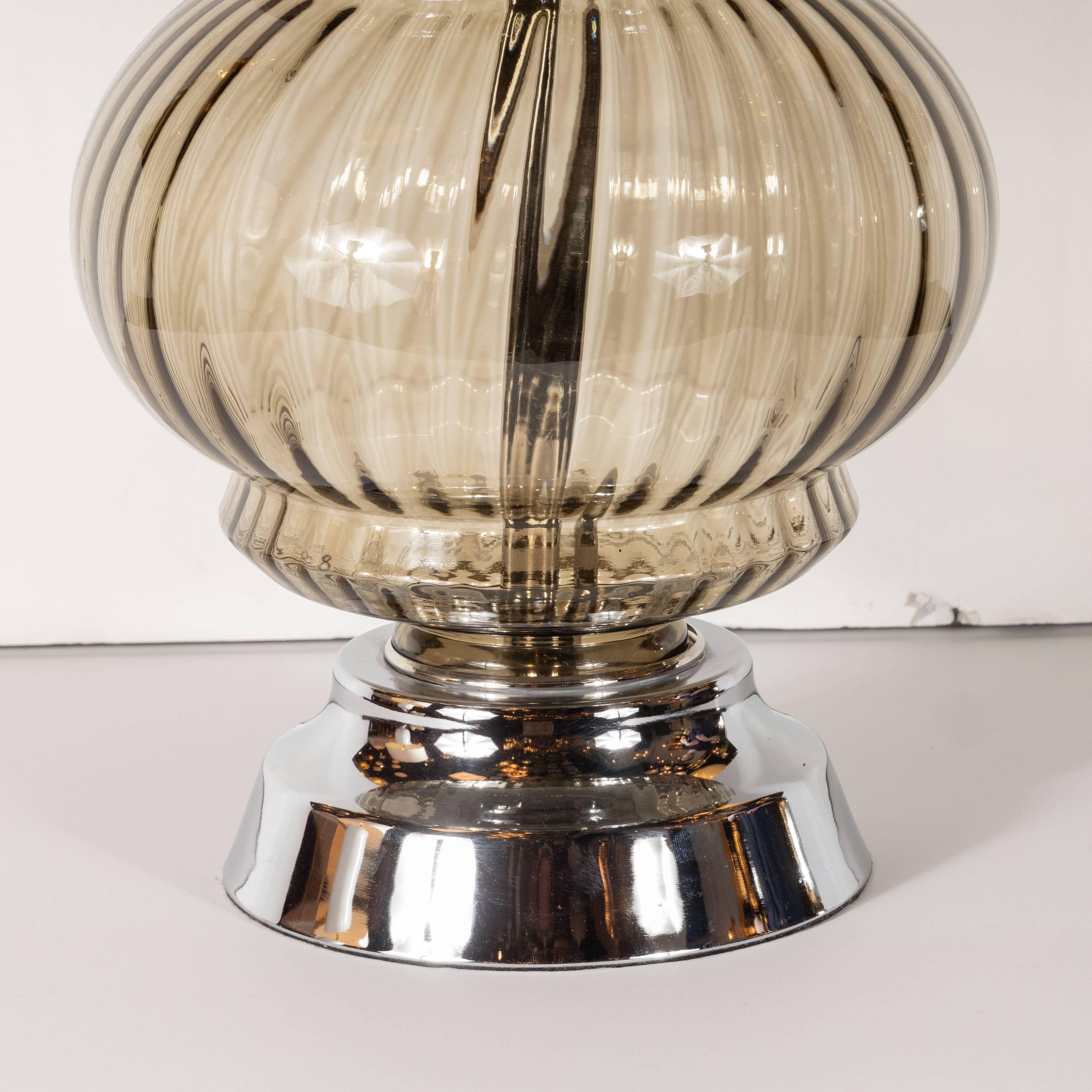 This elegant Mid-Century Modern table lamp was realized in the United States, circa 1960. It features a ribbed orbital glass body with a stepped indentation on top and bottom all in translucent smoked glass. The body sits on a gently sloped and