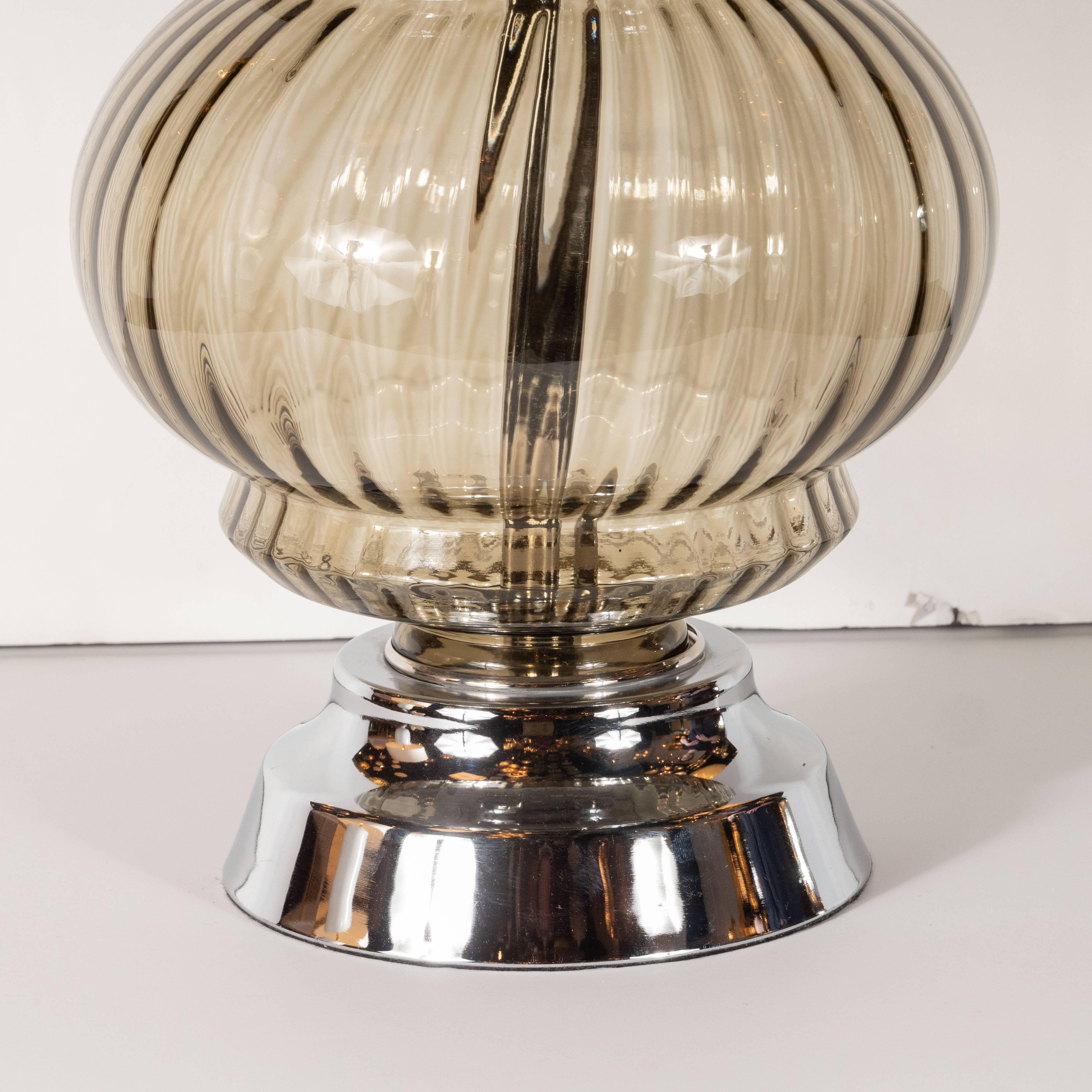 Esta elegante lámpara de mesa Moderna de mediados de siglo se realizó en Estados Unidos, hacia 1960. Presenta un cuerpo de cristal orbital estriado con una hendidura escalonada en la parte superior e inferior, todo en cristal ahumado translúcido. El