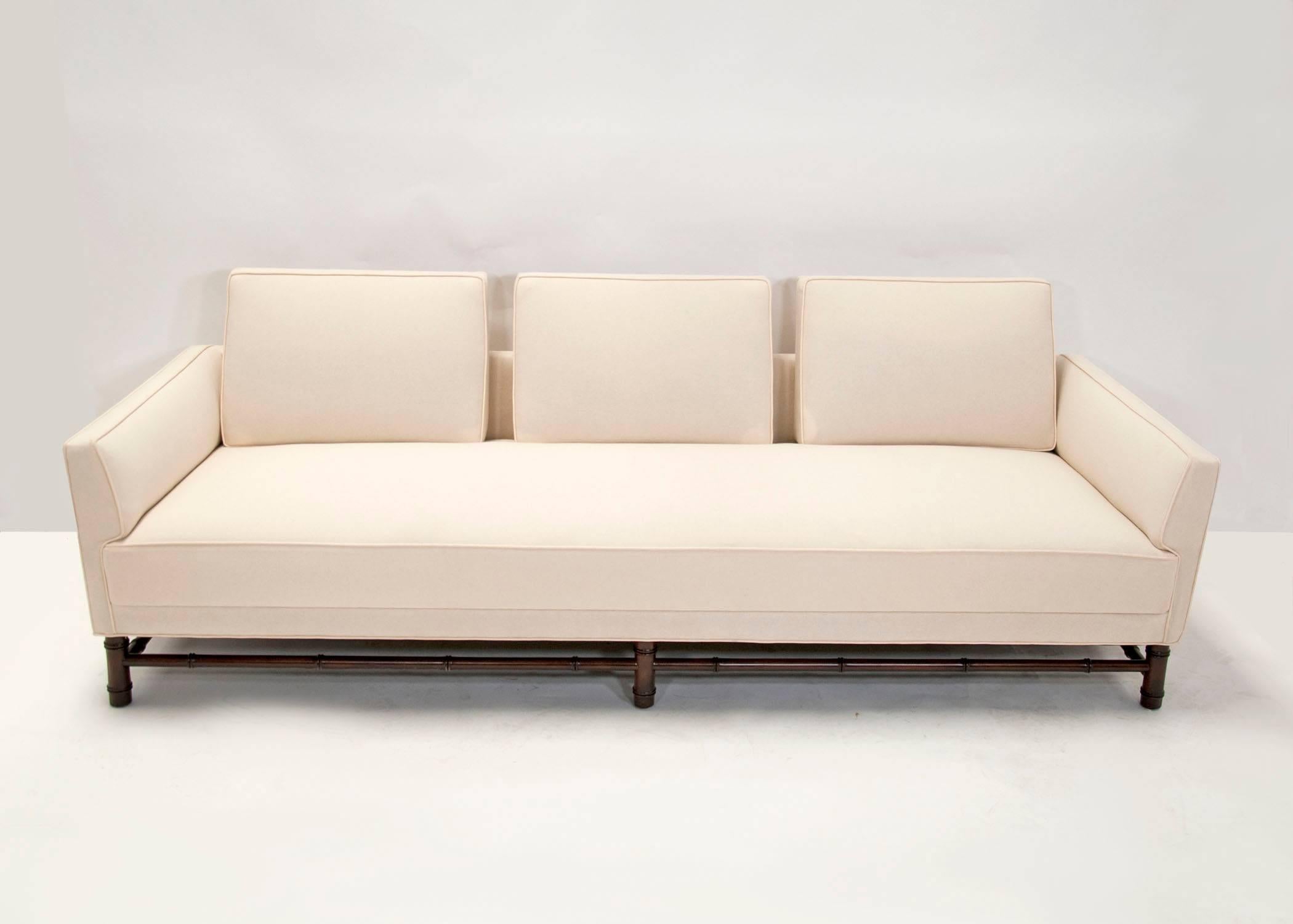A custom Mid-Century Modern sofa in the Hollywood Regency style. Flared arms with a St Thomas style arm popularized by Billy Baldwin. Tight seat with three attached pillow backs on a faux bamboo base. Restored and re-upholstered in of white muslin.