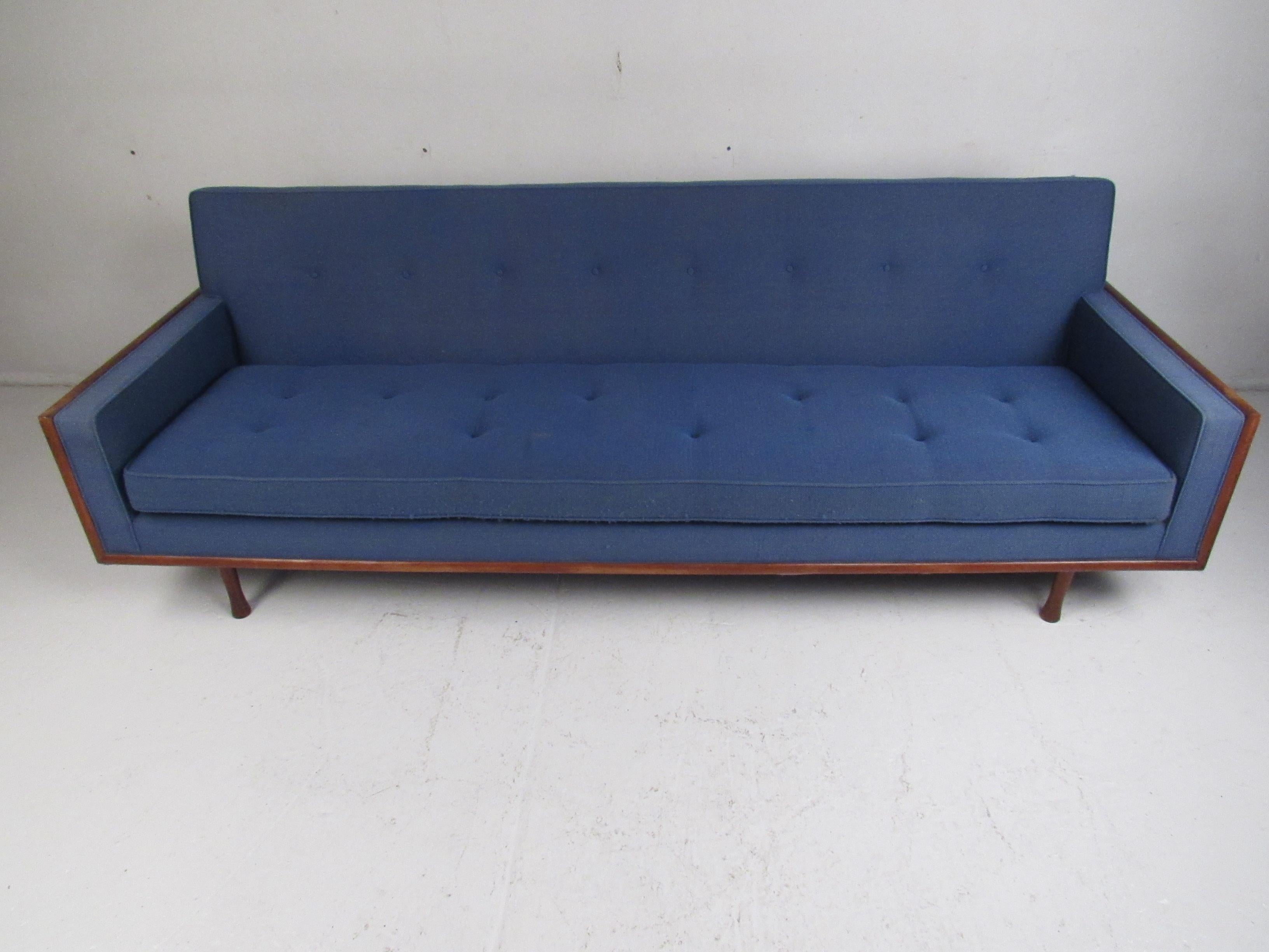 This attractive vintage modern sofa boasts a single overstuffed cushion covered in a plush royal blue upholstery. The unique drum stick shaped legs and walnut trim along the edges add to the allure. A straight line midcentury design with tufted