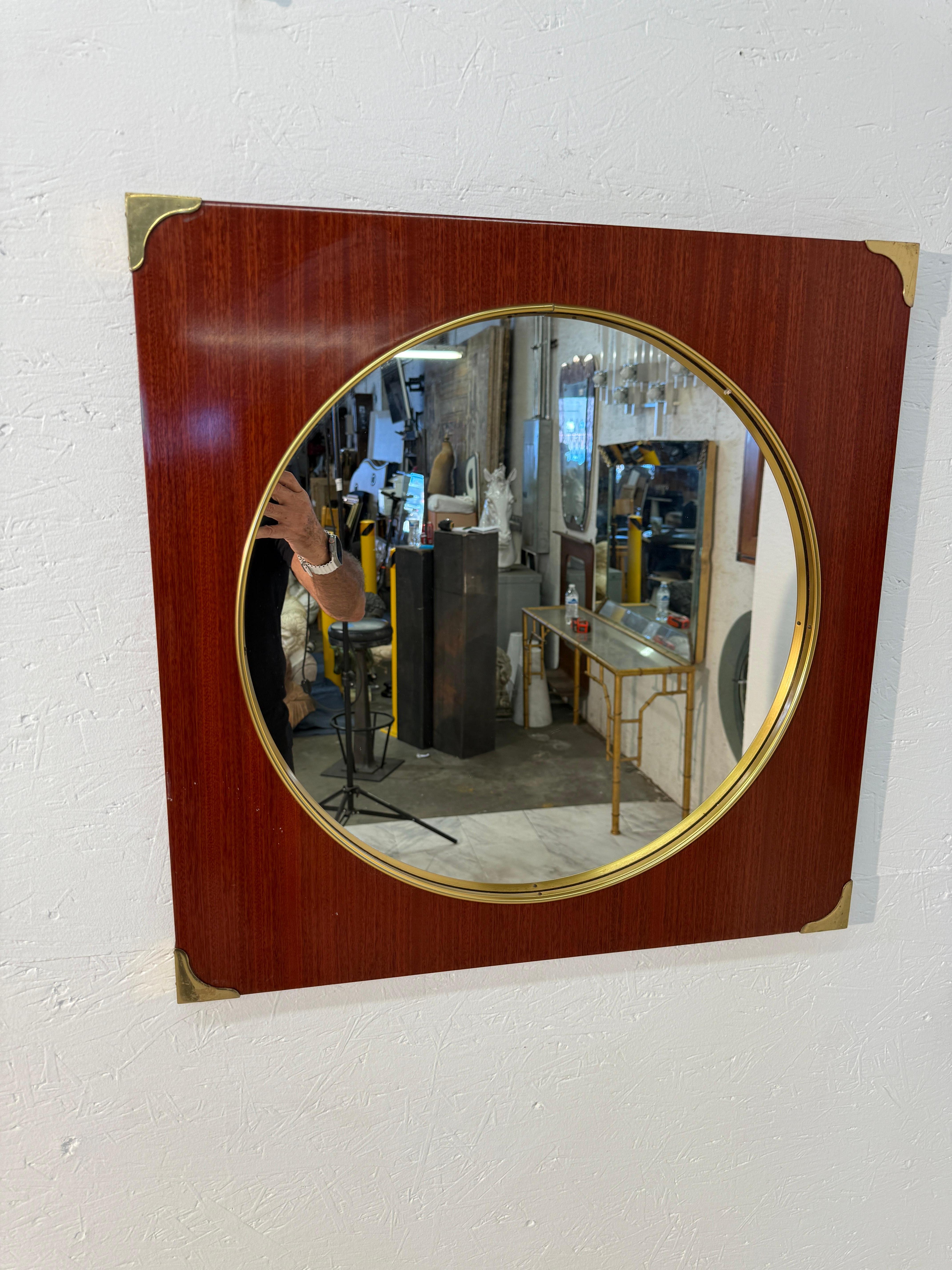 Este elegante espejo de pared moderno de mediados de siglo tiene un panel cuadrado de chapa de madera en un tono rico y cálido, con un espejo circular enmarcado en latón pulido. Cada esquina del panel cuadrado está acentuada con esquineras