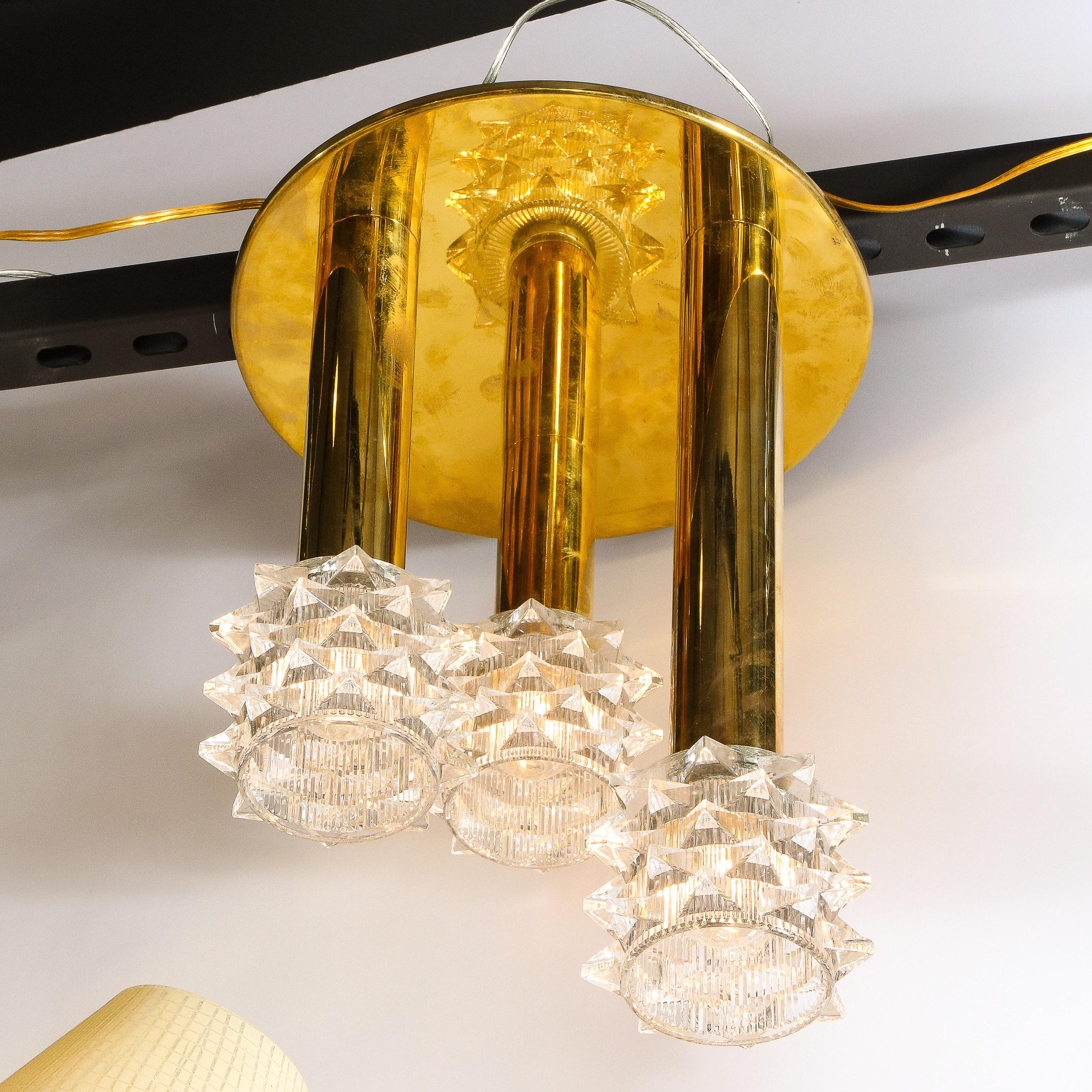 This sophisticated Mid-Century Modern flush mount chandelier was realized in Germany, circa 1970. It features three staggered cylindrical arms that descend from a circular base all in polished brass. The arms attach to cylindrical shades in