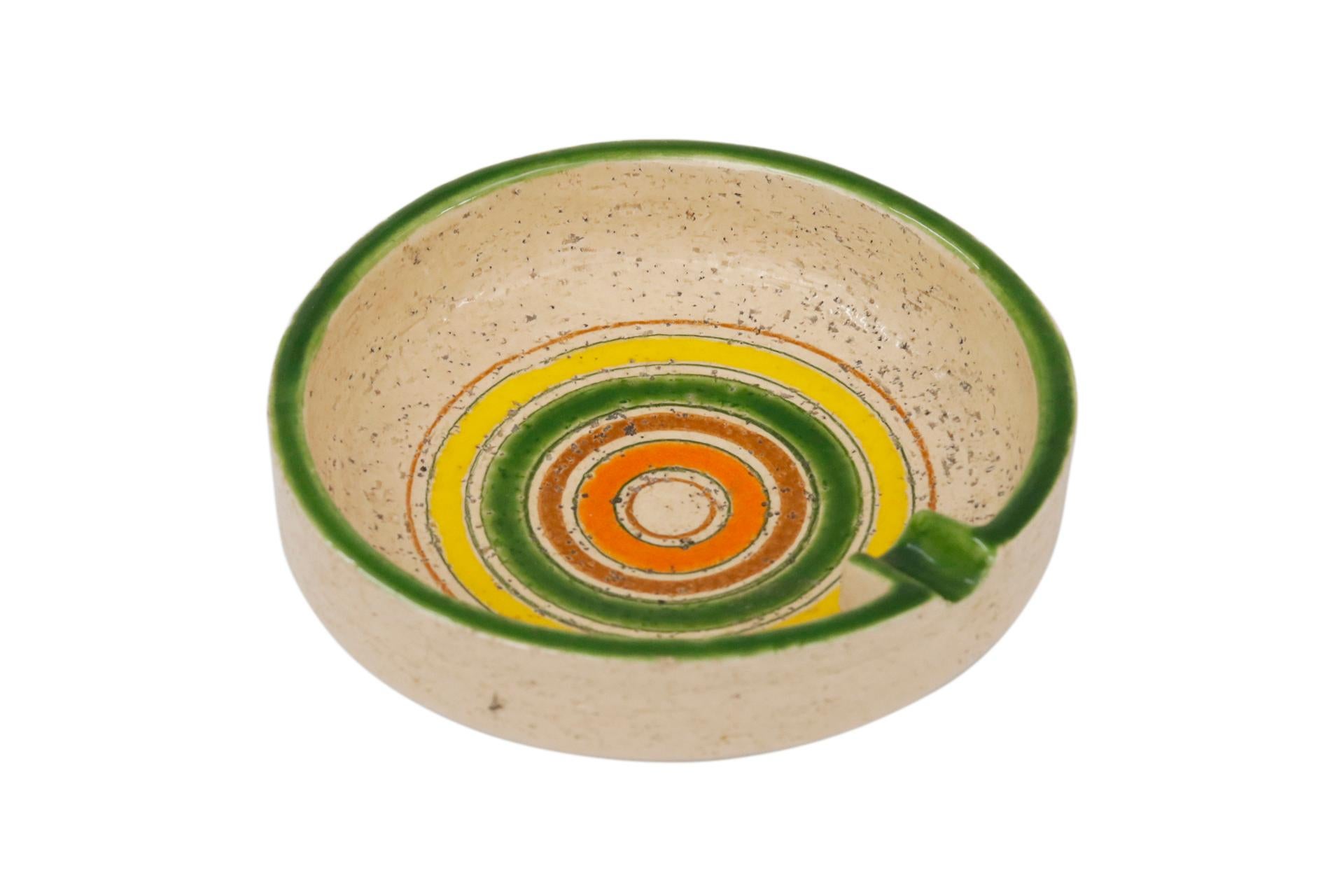 A 1970’s Bitossi Italian ceramic ashtray decorated with colorful concentric circles in orange, brown, green and yellow on a tan background. The rim is finished in green with a single cigarette holder. Marked Italy underneath.
