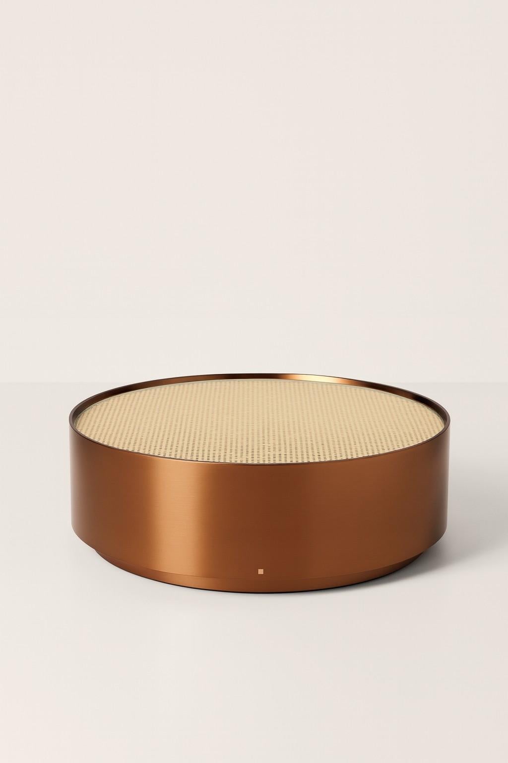 La table centrale en bois massif présente une forme arrondie en finition laquée métallique. Une base encastrée dans le même bois assure un aspect cohérent, tandis que le plateau de table scelle le rotin tressé avec du verre extra-clair, créant ainsi