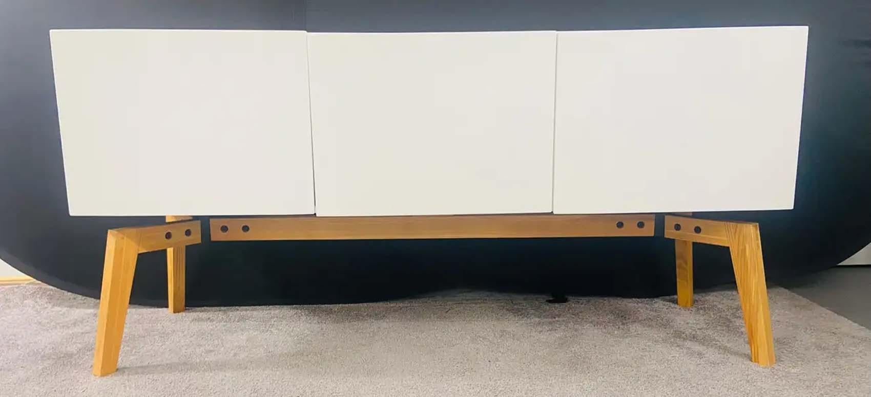 A stylish Mid- Century Modern credenza, sideboard or cabinet featuring three doors opening to reveal plenty of storage space. The credenza is made in lacquered white finish and is resting on four tapered and angled wooden legs. Simple, yet elegant