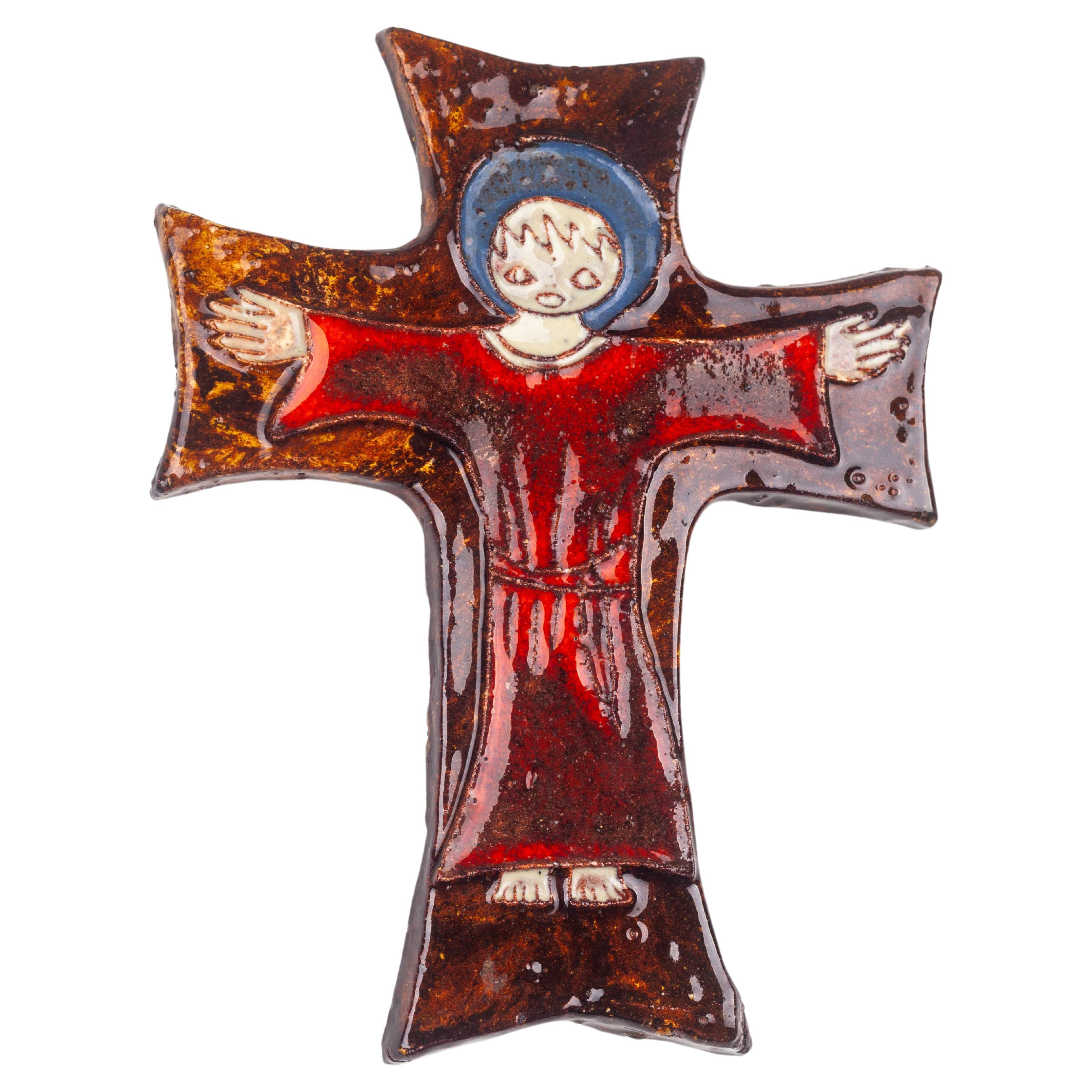 This ceramic cross, handcrafted by studio pottery artists in mid-century Europe, presents a stylized figurative scene within its bounds, evoking the rich tradition of religious art reimagined through the lens of modern design. The figure, likely