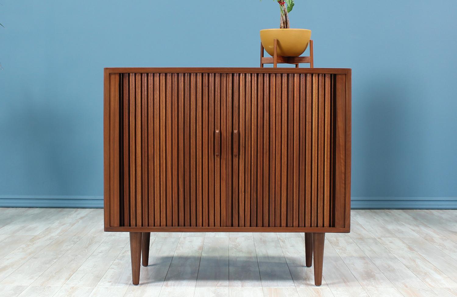 Mid-Century Modern Cabinet designed and manufactured in the United States circa 1950’s. This compact and practical cabinet features a walnut wood case that sits over tapered legs. The carved pulls on the tambour doors allow them to glide open