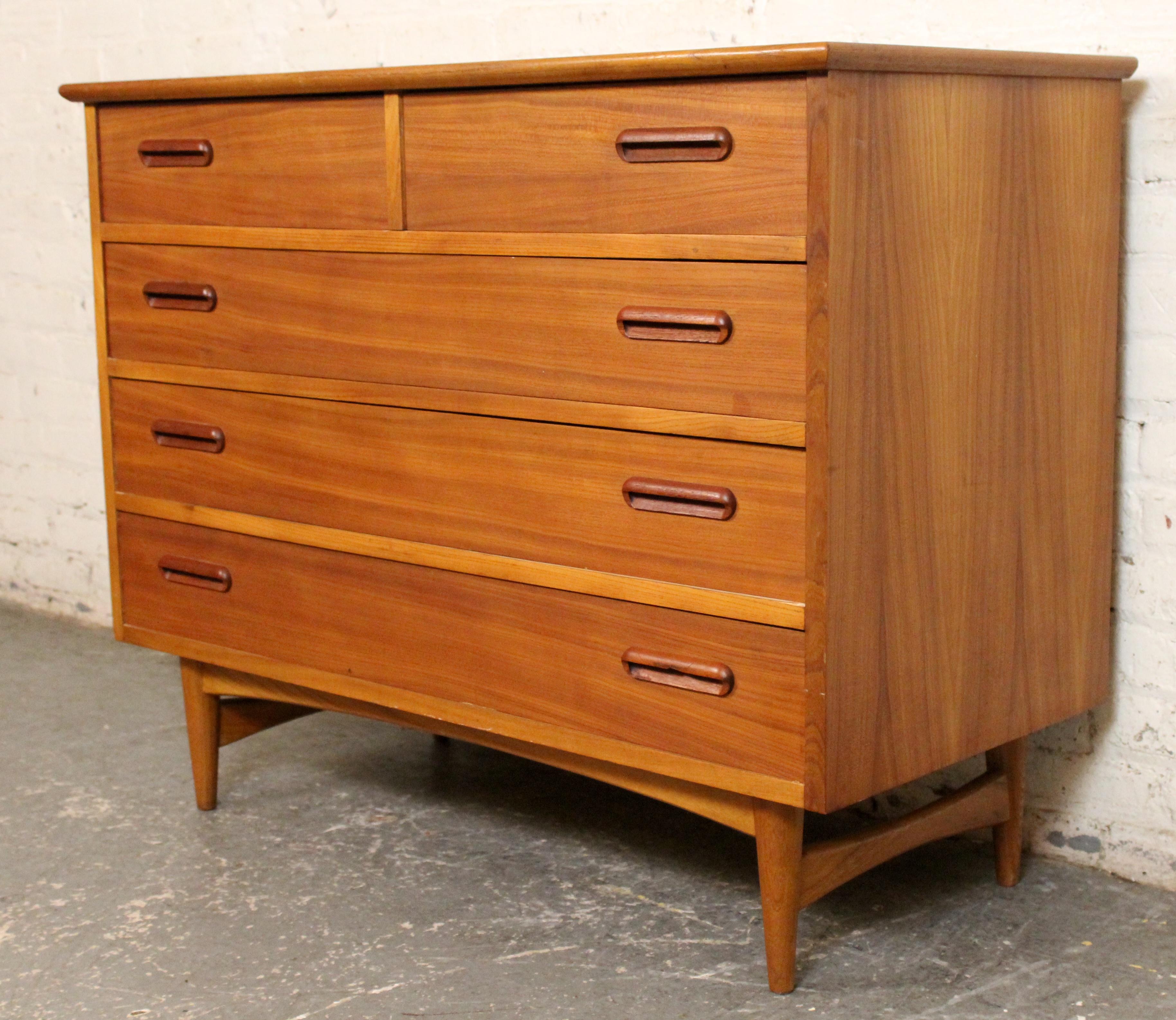 A striking example of Mid-Century Modern design, this teak chest of drawers features clean lines, warm wood grain, and sculpted recessed handles. Comprising five drawers—including two smaller ones at the top—this piece offers ample storage while
