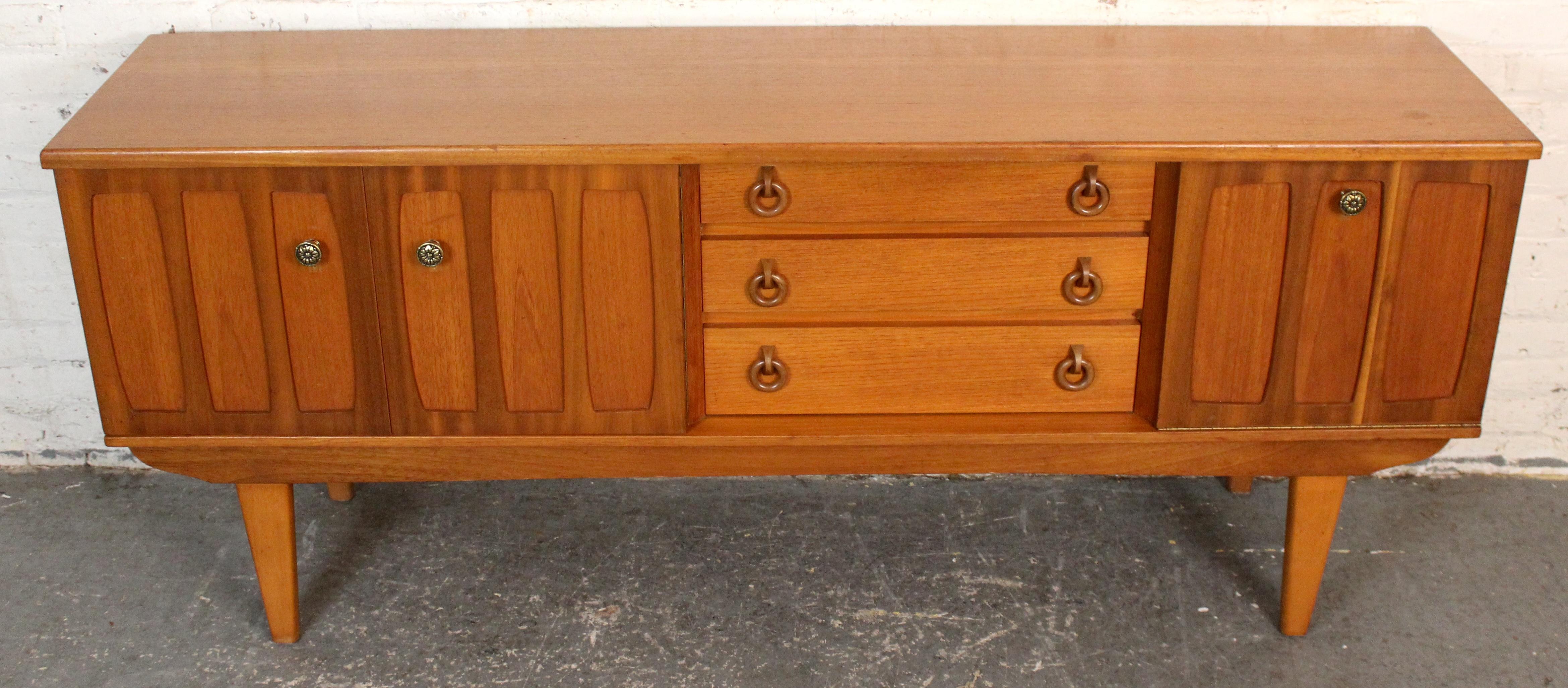 Esempio di artigianato britannico di metà secolo, questa credenza di Wood Beautility presenta linee pulite e calde tonalità di legno di teak. Il pezzo presenta un design simmetrico con tre cassetti centrali affiancati da due armadietti, ciascuno