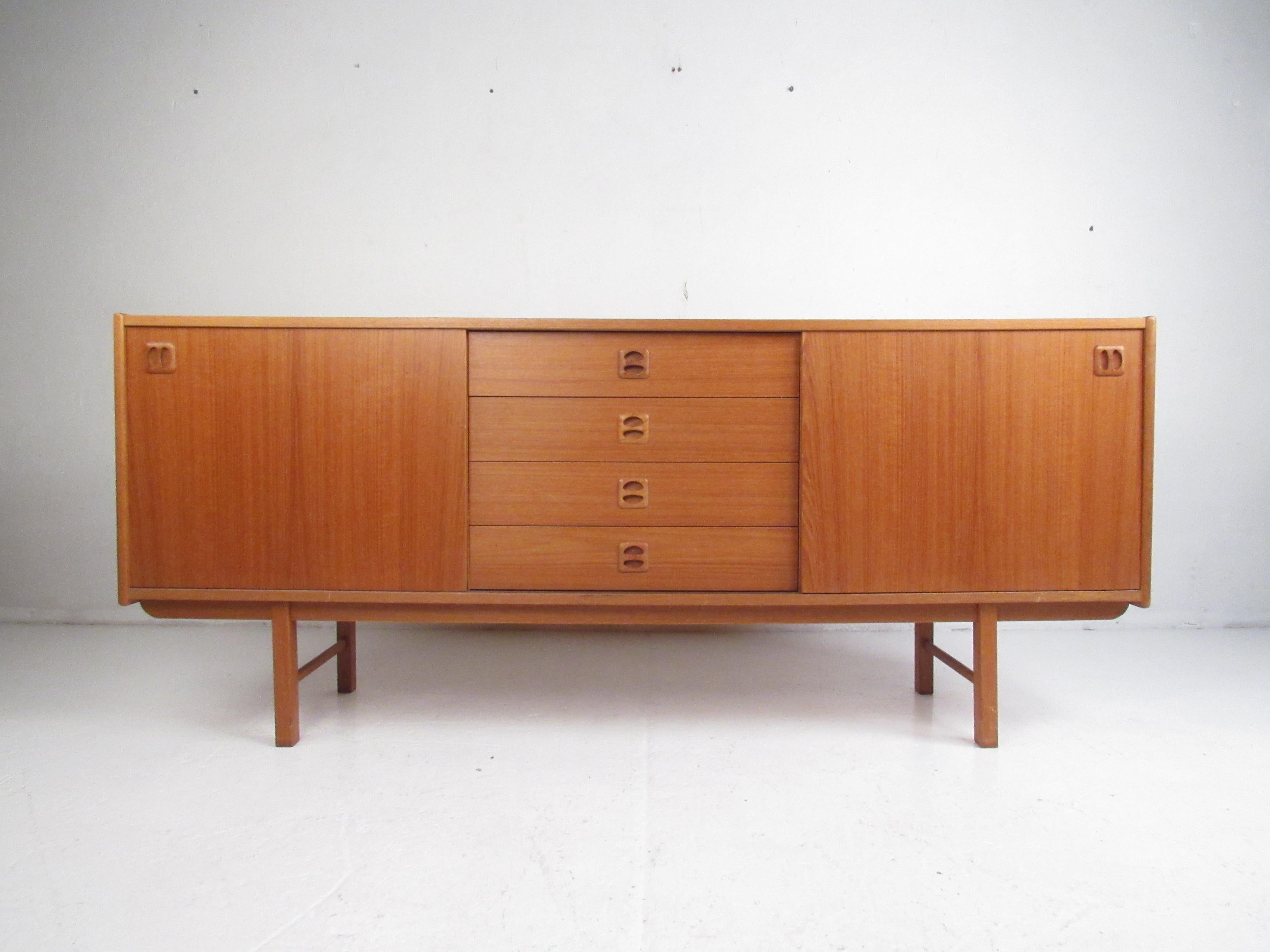 This beautiful vintage modern sideboard features two large compartments hidden by sliding doors and four hefty drawers in the center. An attractive Danish design with carved ellipse shaped pulls, a rich teak finish, and drawers with dovetailed
